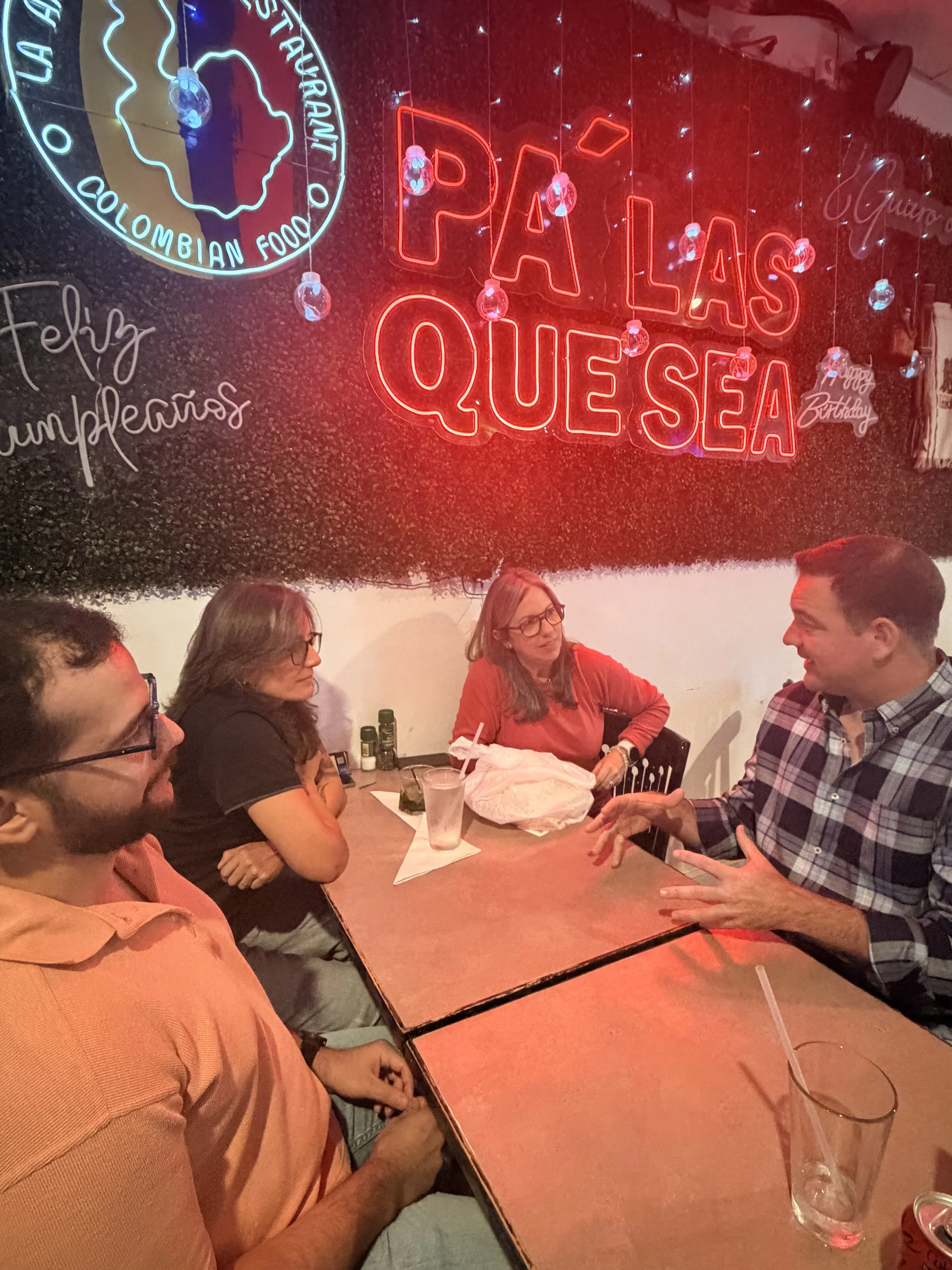 Group of four people sitting at a restaurant table, engaged in conversation. Neon signs in the background read 'PÁ LAS QUE SEA' and 'Colombian Food', with decorative lighting and a person wearing glasses visible.