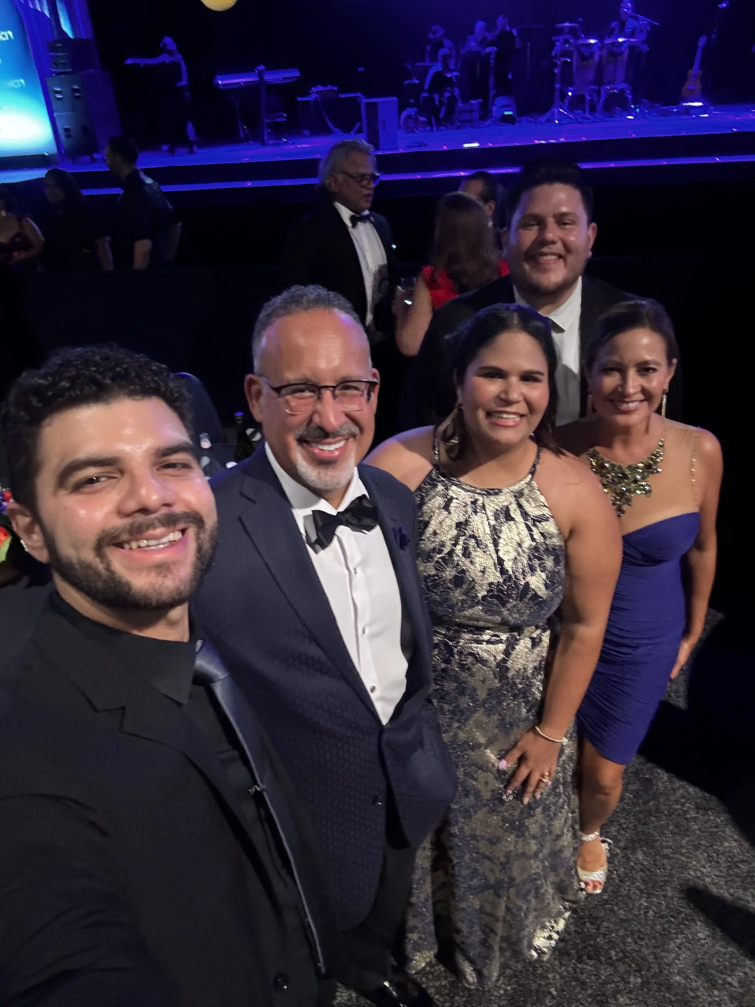 Group of five people in formal attire taking a selfie at a fancy event with live band performing on stage in the background.