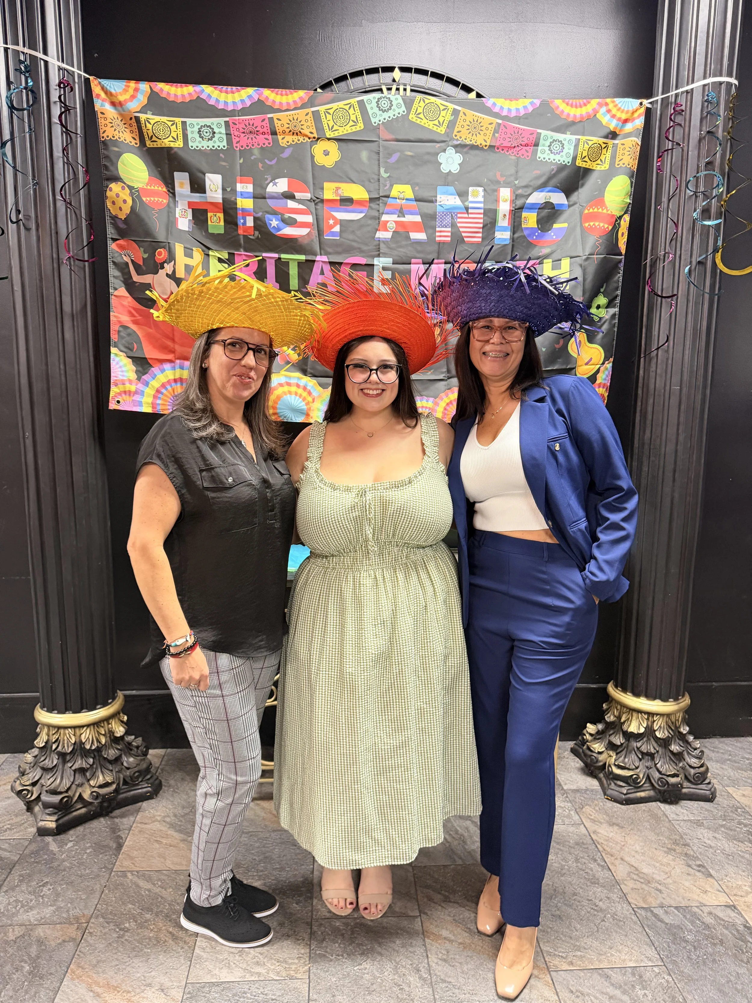 Three women wearing colorful sombreros standing in front of a 'Hispanic Heritage' banner at a celebration event.