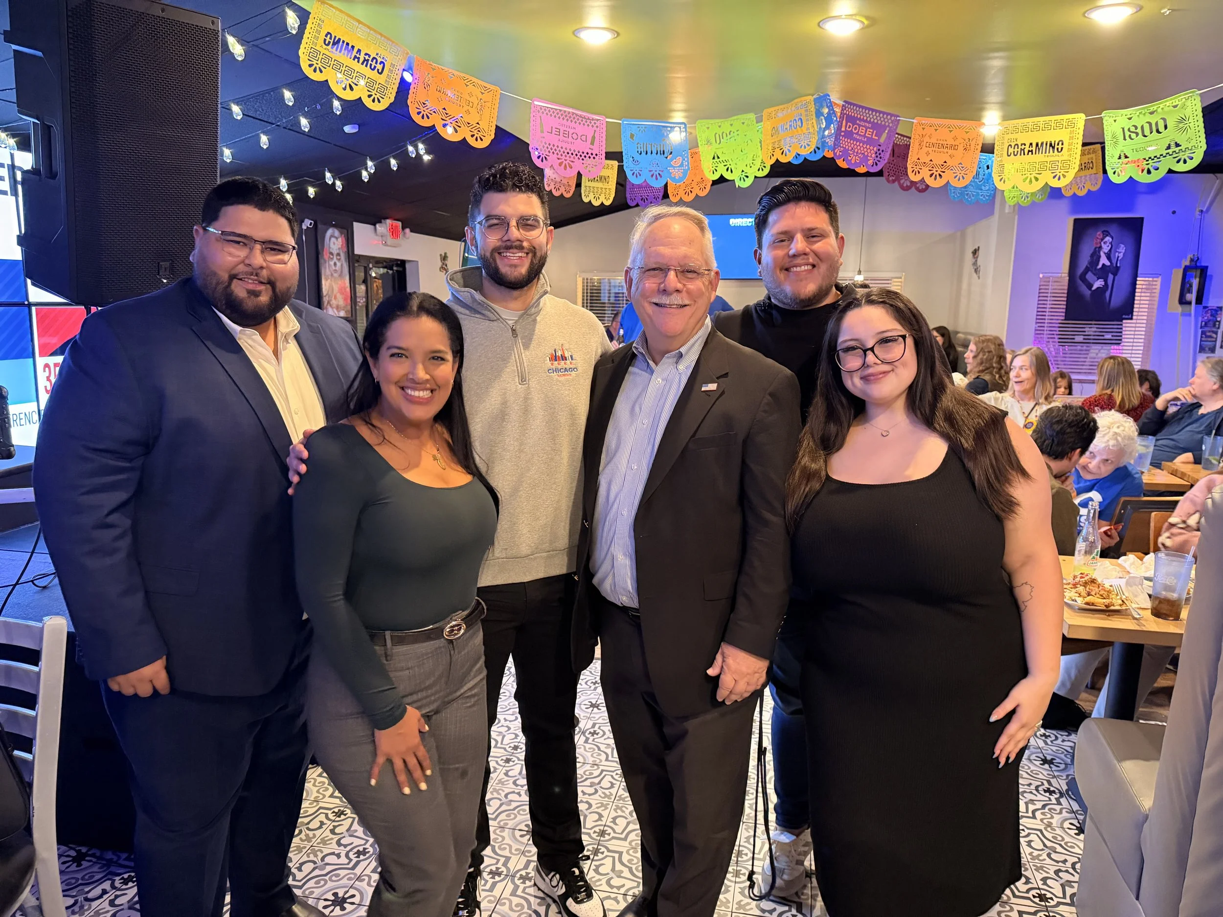 Group of six people smiling at a celebration in a decorated indoor venue, with colorful papel picado banners hanging overhead and guests seated in the background.