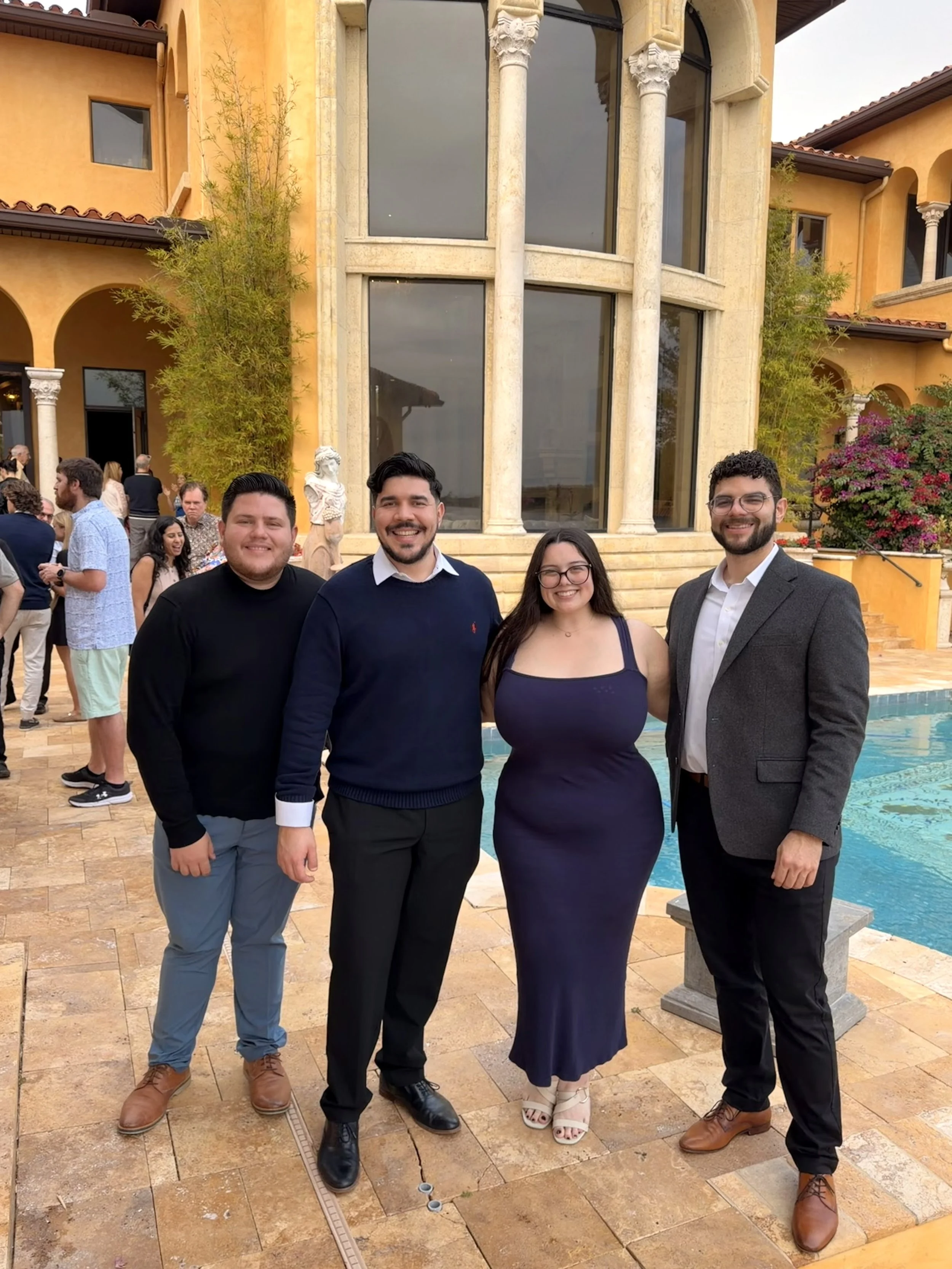Four young adults, two men and one woman, standing outside near a swimming pool with a large, ornate beige building in the background. They are dressed nicely, smiling for the photo, at what appears to be a social gathering or celebration.