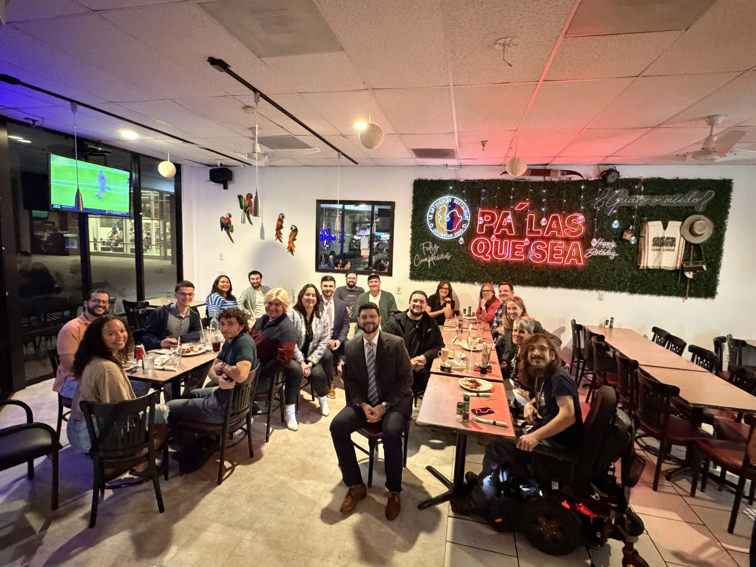 A group of people celebrating a birthday at a restaurant with festive decorations, neon signs, and a large window showing the street outside.
