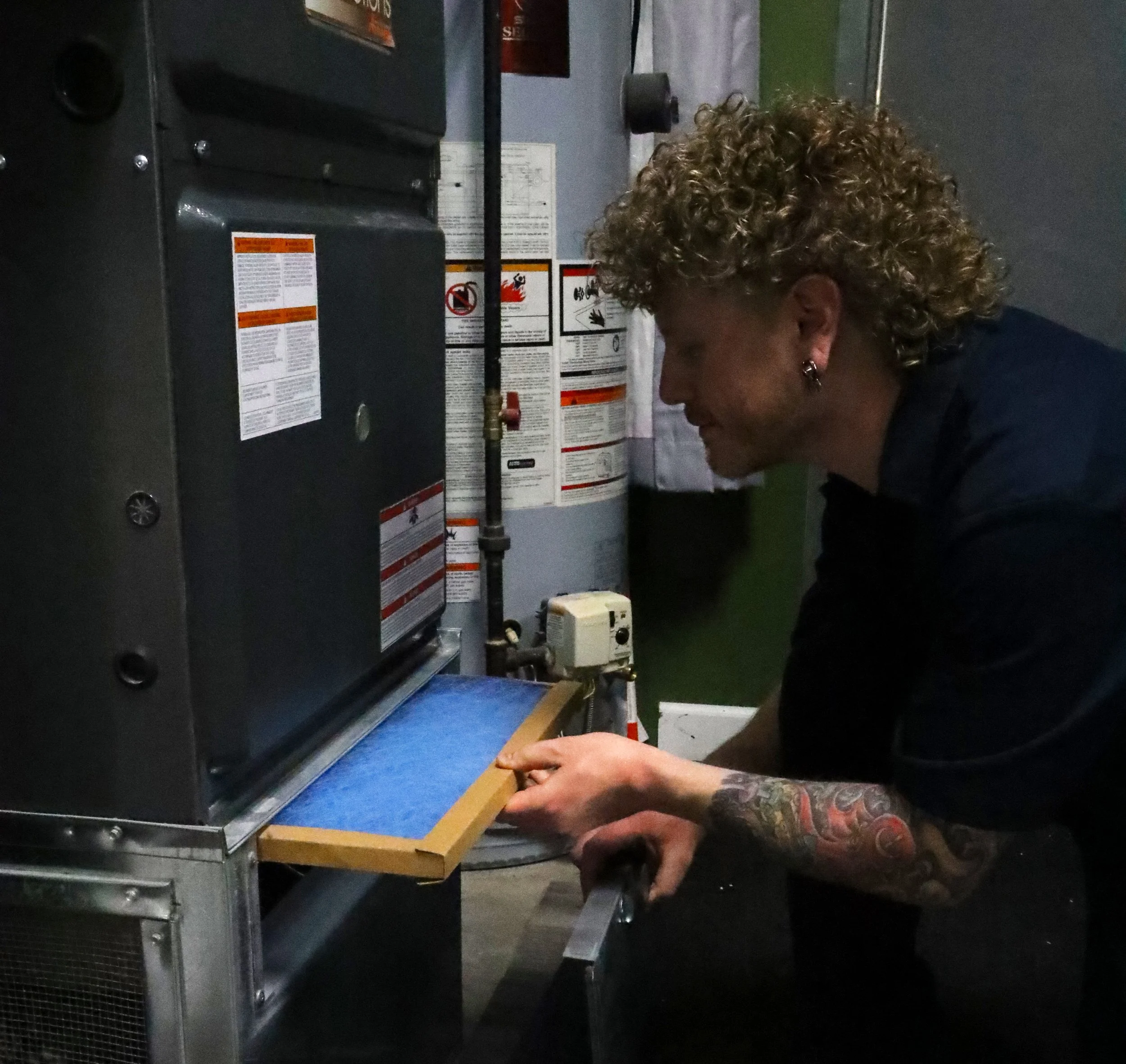 A person with curly hair and tattoos on their arm is working with a piece of equipment that appears to be an industrial appliance or machine, with various warning labels and pipes attached, in a room with industrial and safety components.