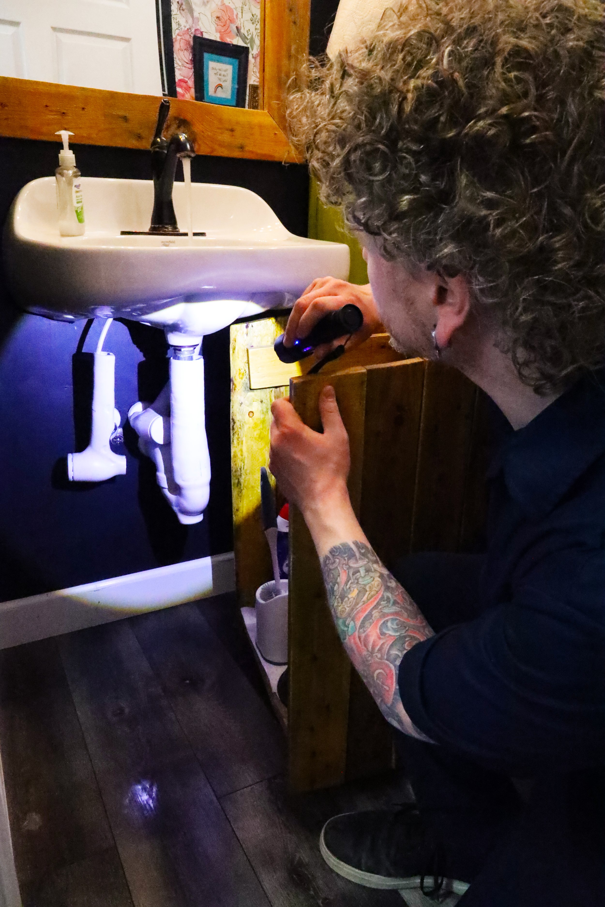 Person with curly hair and colorful tattoos crouched using a flashlight to inspect a small wall-mounted white sink with black fixtures, in a room with dark wooden flooring, a mirror, and framed artwork on the wall.