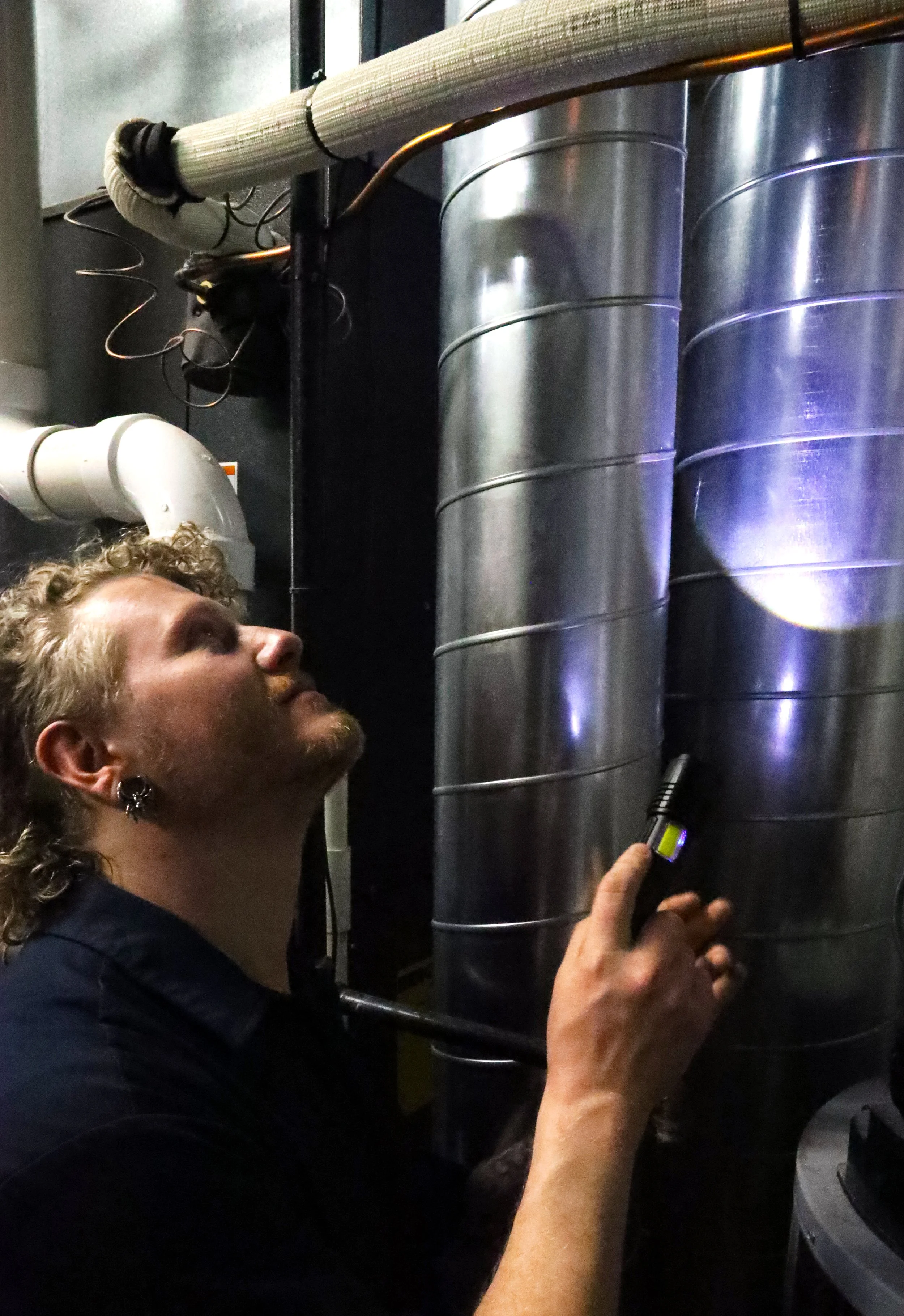 A man with curly hair and a beard inspecting industrial equipment with a flashlight, standing next to large metallic cylinders and pipes.