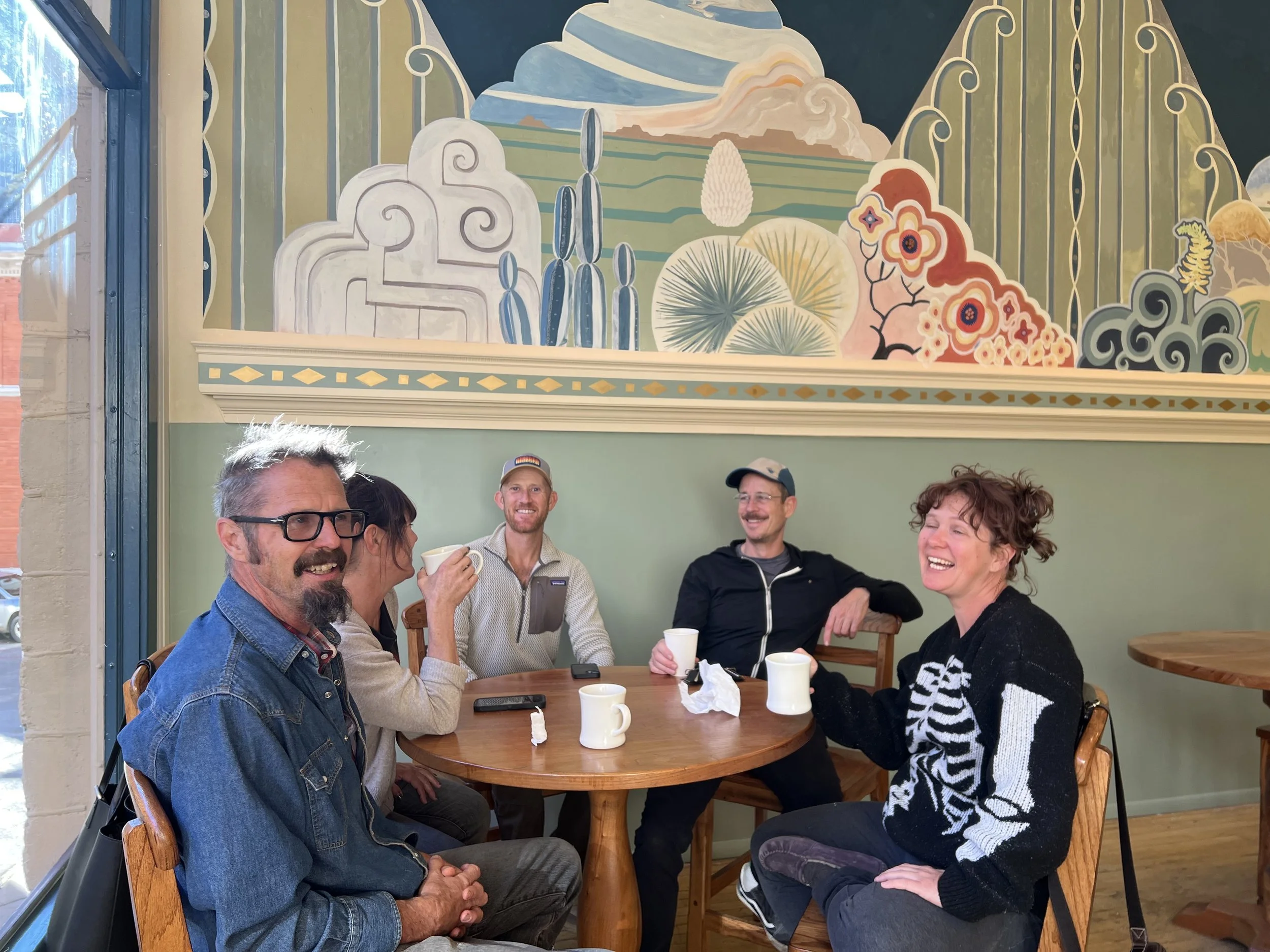 Five people sitting around a wooden table in a cafe, smiling and conversing. The cafe has a colorful mural on the wall behind them, featuring stylized trees, clouds, and geometric patterns.