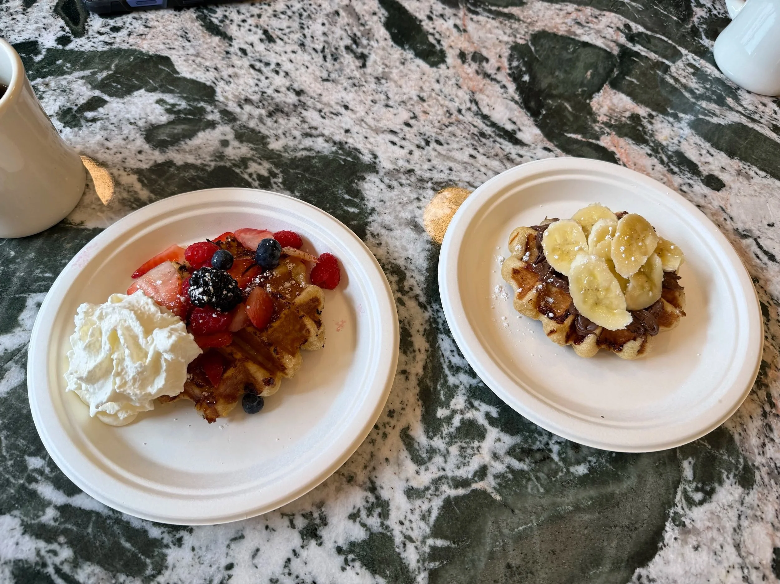 Waffles topped with bananas and chocolate, served on white plates, with additional whipped cream and fresh berries on one waffle.