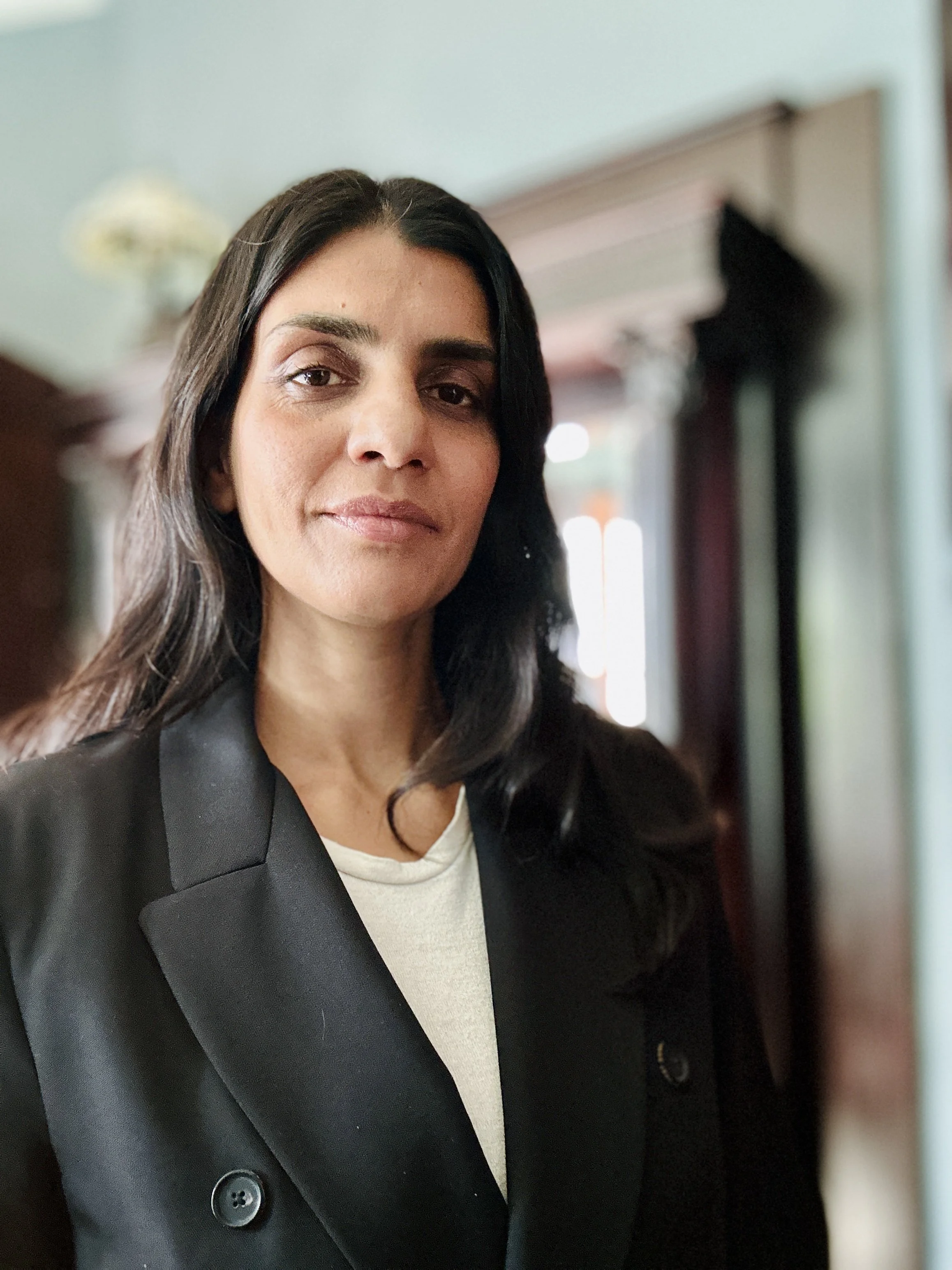 A woman with dark hair, wearing a black blazer and white top, standing indoors with a blurred background