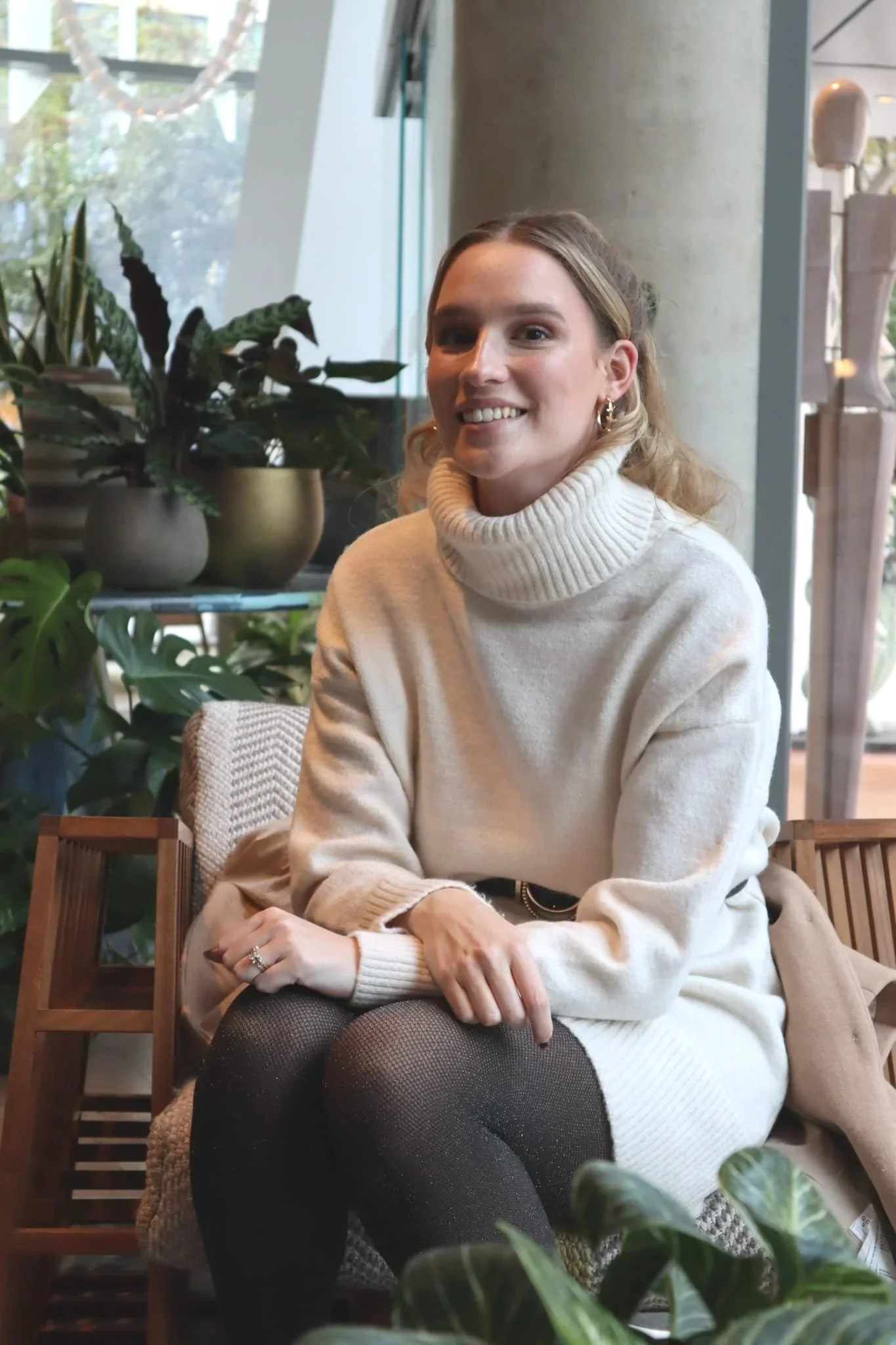 A smiling woman with long, blonde hair, wearing a cream turtleneck sweater and black textured tights, sitting in a cozy, plant-filled indoor space.