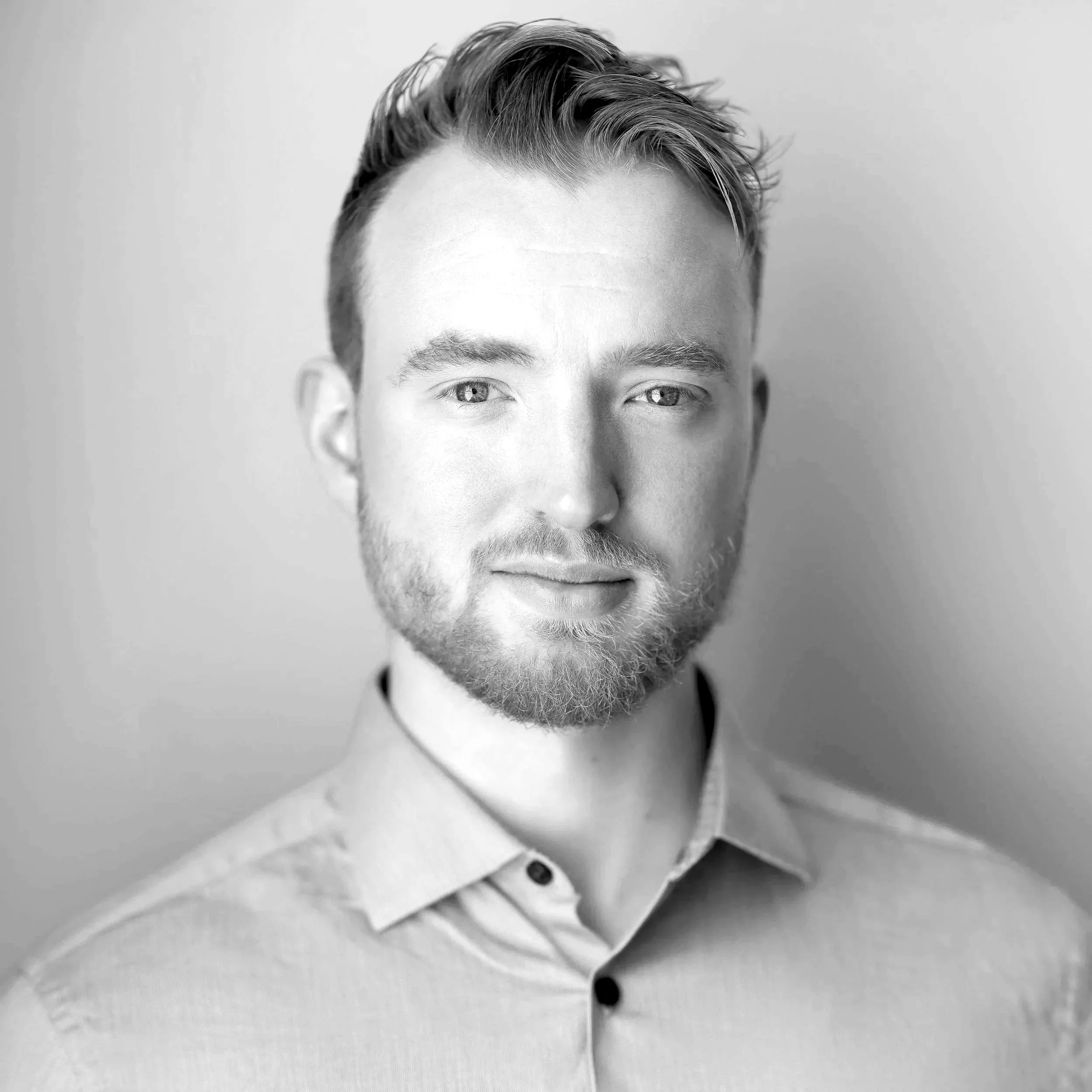 Black and white portrait of a young man with short, styled hair and a beard, wearing a collared shirt, smiling softly at the camera.