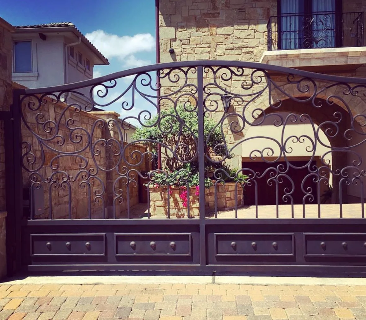 Decorative black wrought iron gate with intricate swirls and patterns, leading to a courtyard with stone walls, plants, and a building in the background.