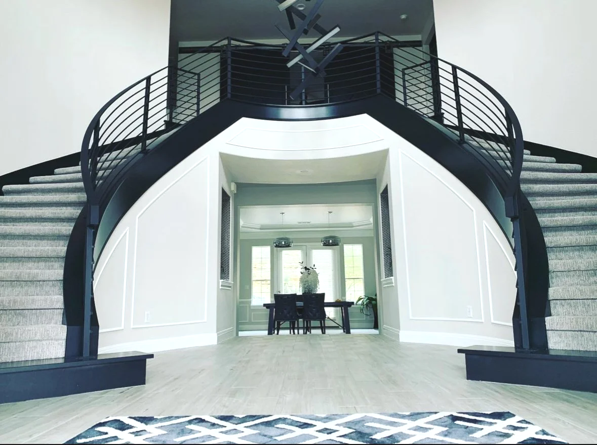 Interior view of a modern home featuring a grand staircase with black railings, leading to a second floor. Behind the staircase, there is a dining area with a table, chairs, a vase with flowers, and large windows.