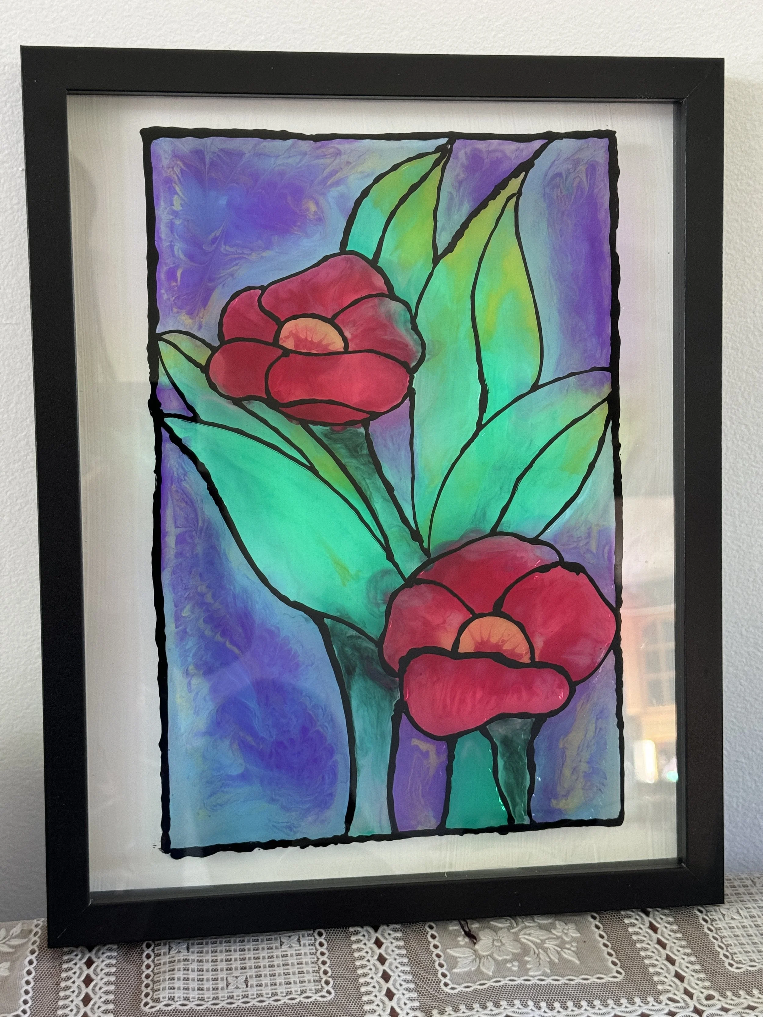 Framed stained glass artwork of two red flowers with orange centers and green leaves, set against a colorful purple and blue background.