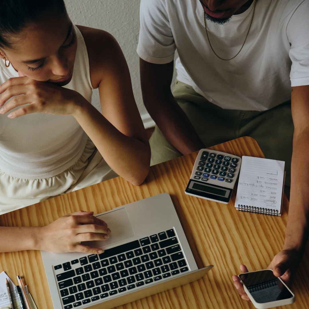 Two people sitting at a wooden table with a laptop, calculator, notepad, pens, and a smartphone, looking at the laptop screen and discussing.