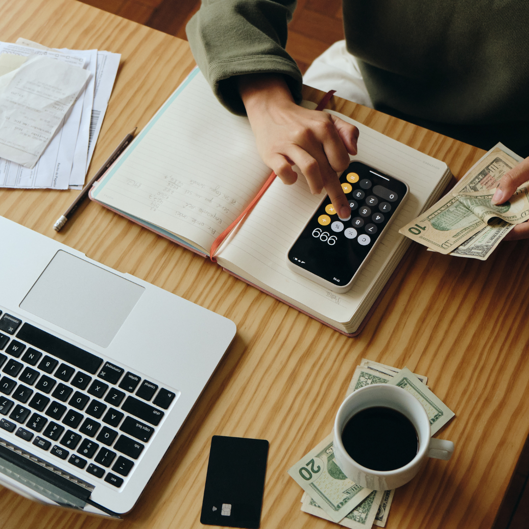 Person using a calculator on a notebook with cash and documents on a wooden desk, alongside a laptop, credit card, a cup of coffee, and a smartphone.