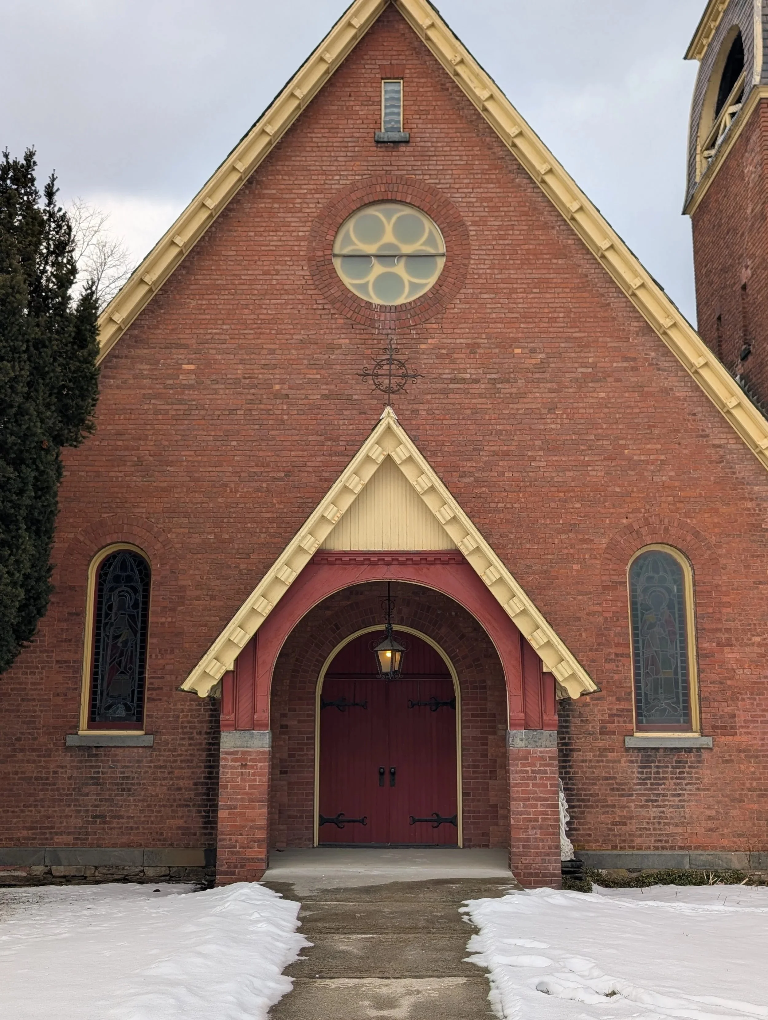 Front view of a red brick church with a pointed roof, stained glass windows, and a wooden door, with snow on the ground.