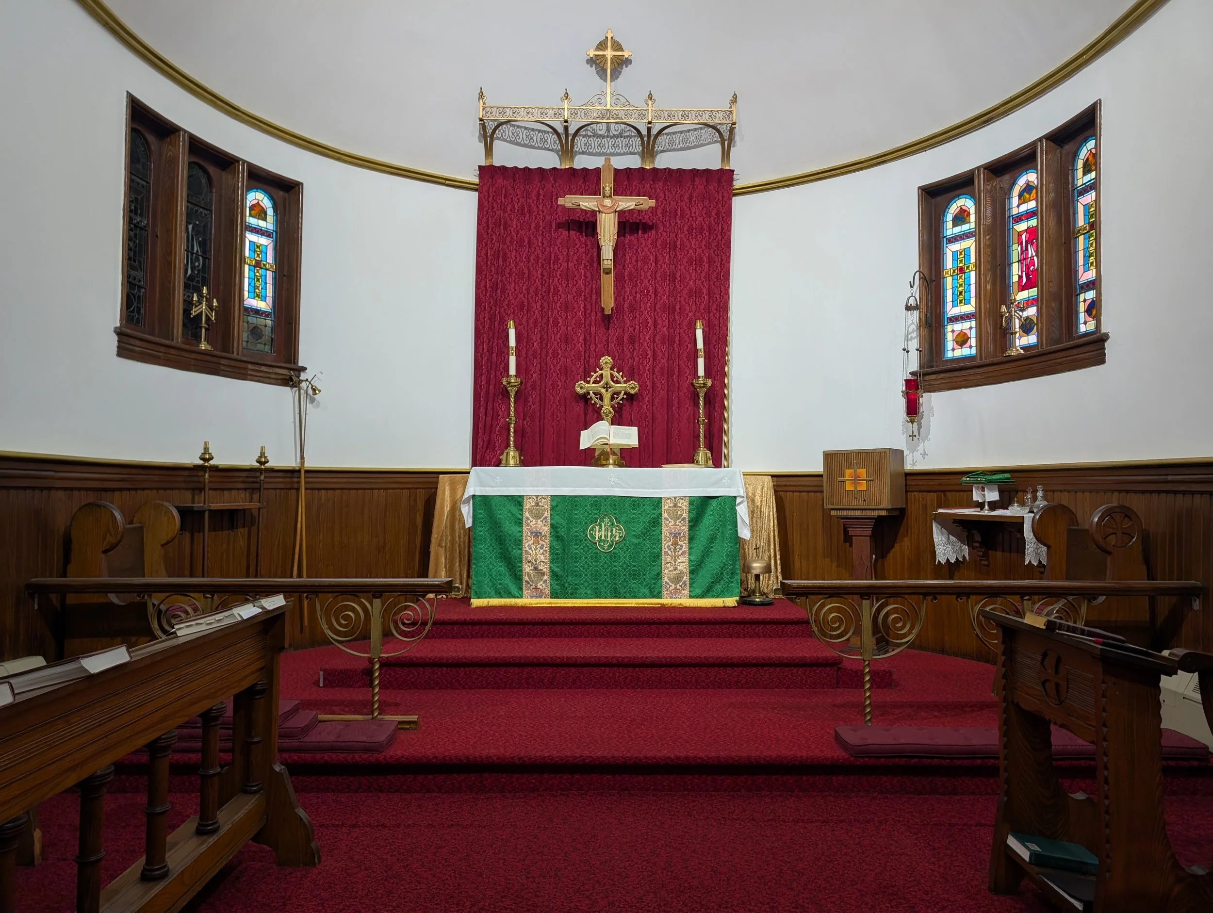 Inside a church altar with red carpeted steps, a crucifix on a red velvet backdrop, gold candle holders, an open book, stained glass windows, and wooden pews.