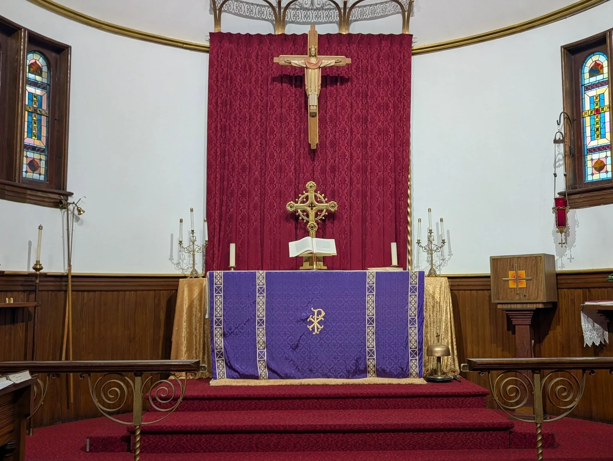 Interior of a church altar with a crucifix above it, draped in a red curtain. The altar has a purple cloth with gold accents, an open Bible, and candelabras. Stained glass windows are on either side.