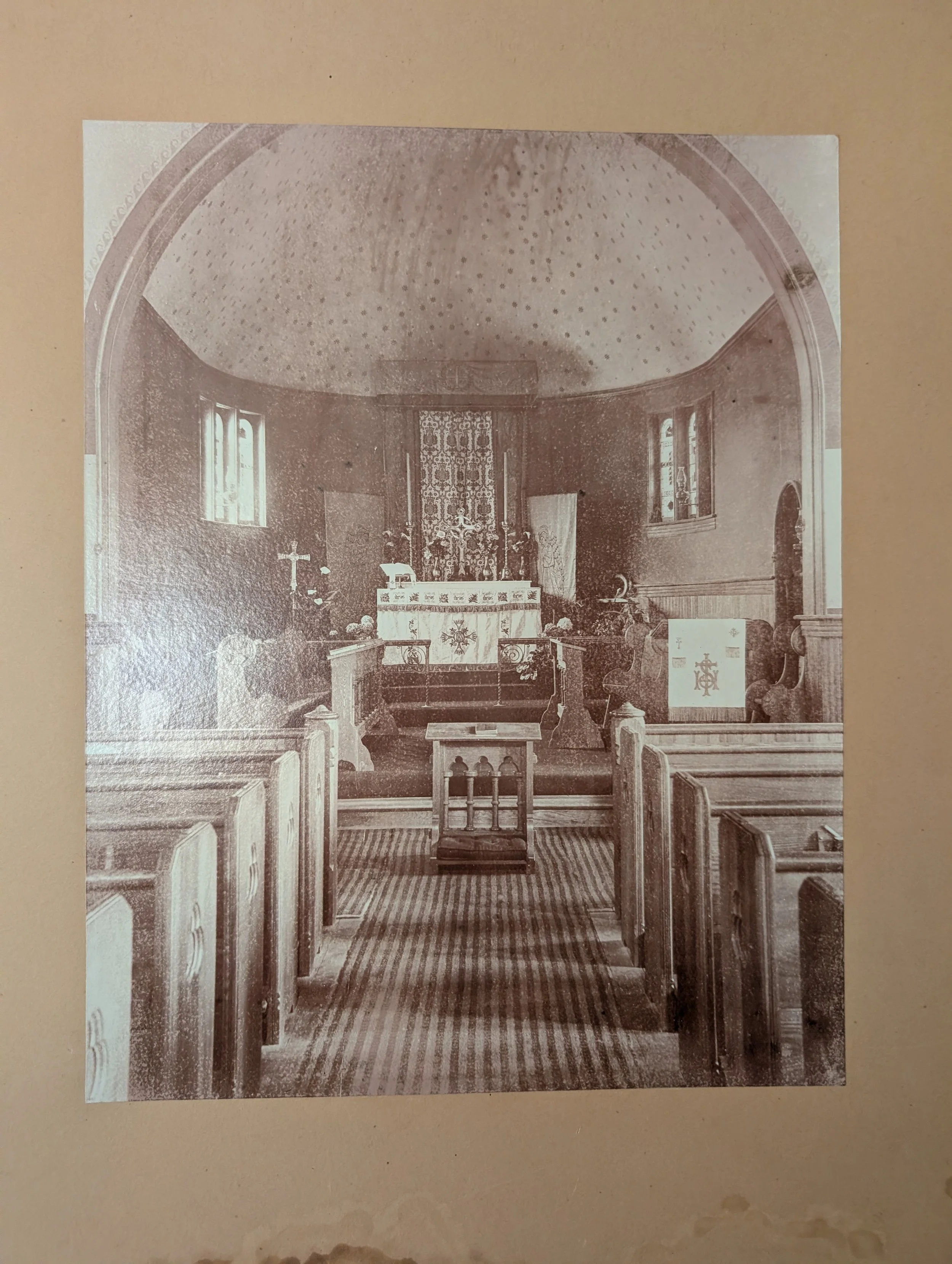 Black and white photo of a small church interior with pews, altar, Latin crosses, and religious symbols, with windows on either side.