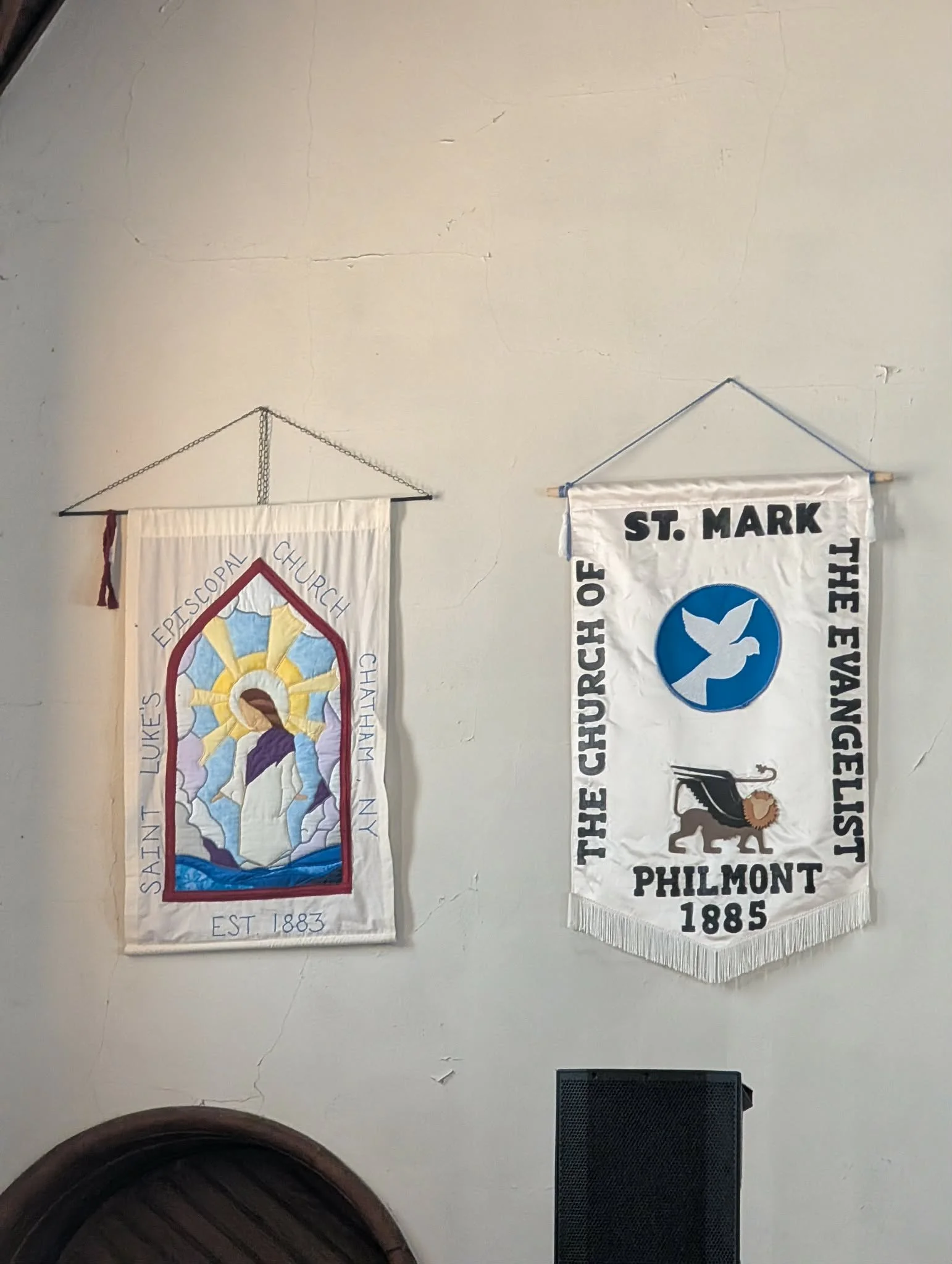 Two banners hanging on a wall inside a church. The left banner depicts a stained glass image of Jesus with a halo, a purple robe, and a white garment, with the text "Saint Luke's Episcopal Church, Chatham N.Y., Est 1883". The right banner features a 