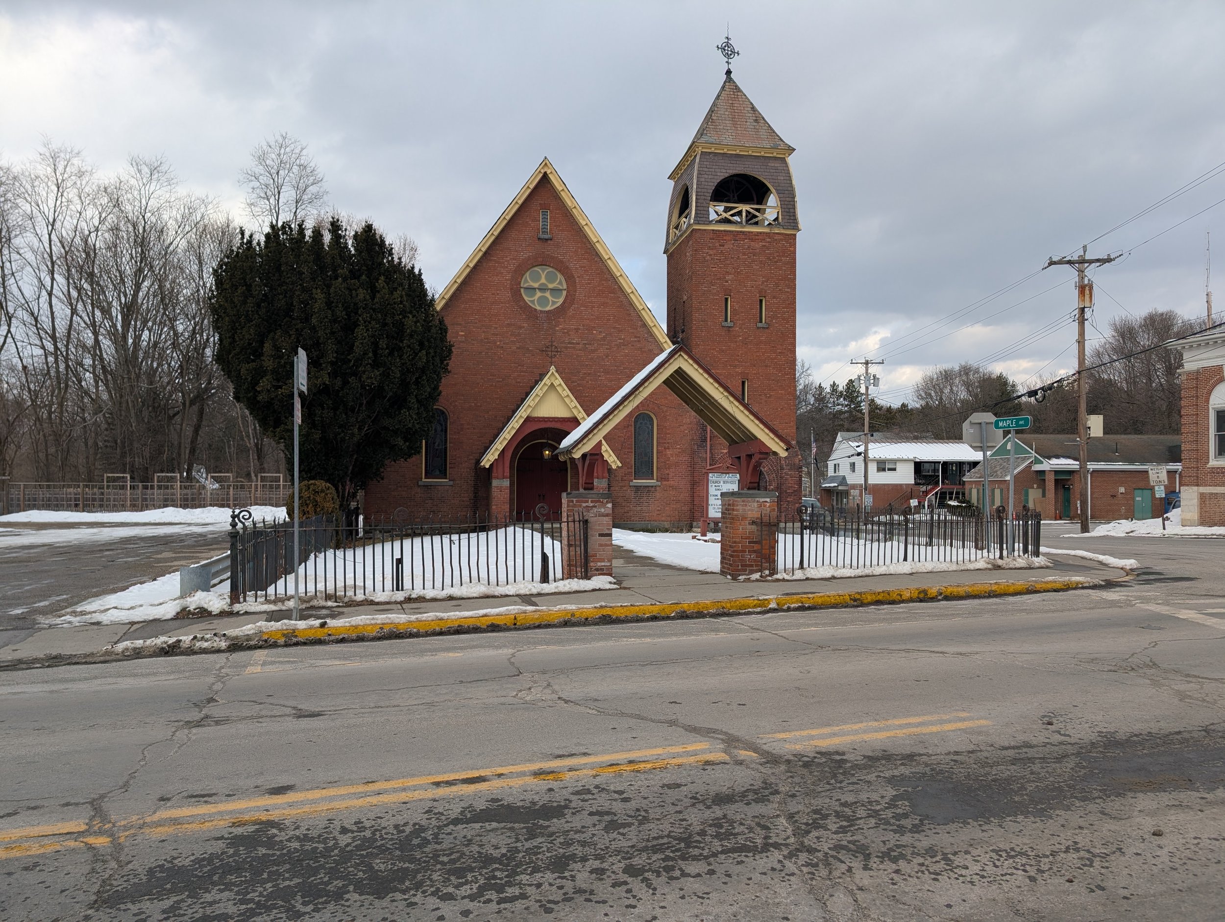 A red brick church with a tall steeple, a large tree to the left, and a small fenced yard in front. Snow is on the ground and the street, with cloudy skies overhead.