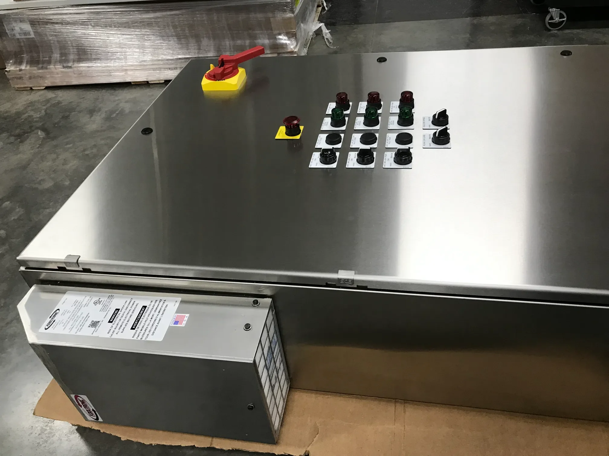 A stainless steel industrial control panel with various switches, knobs, and indicator lights on top, situated in a warehouse setting.