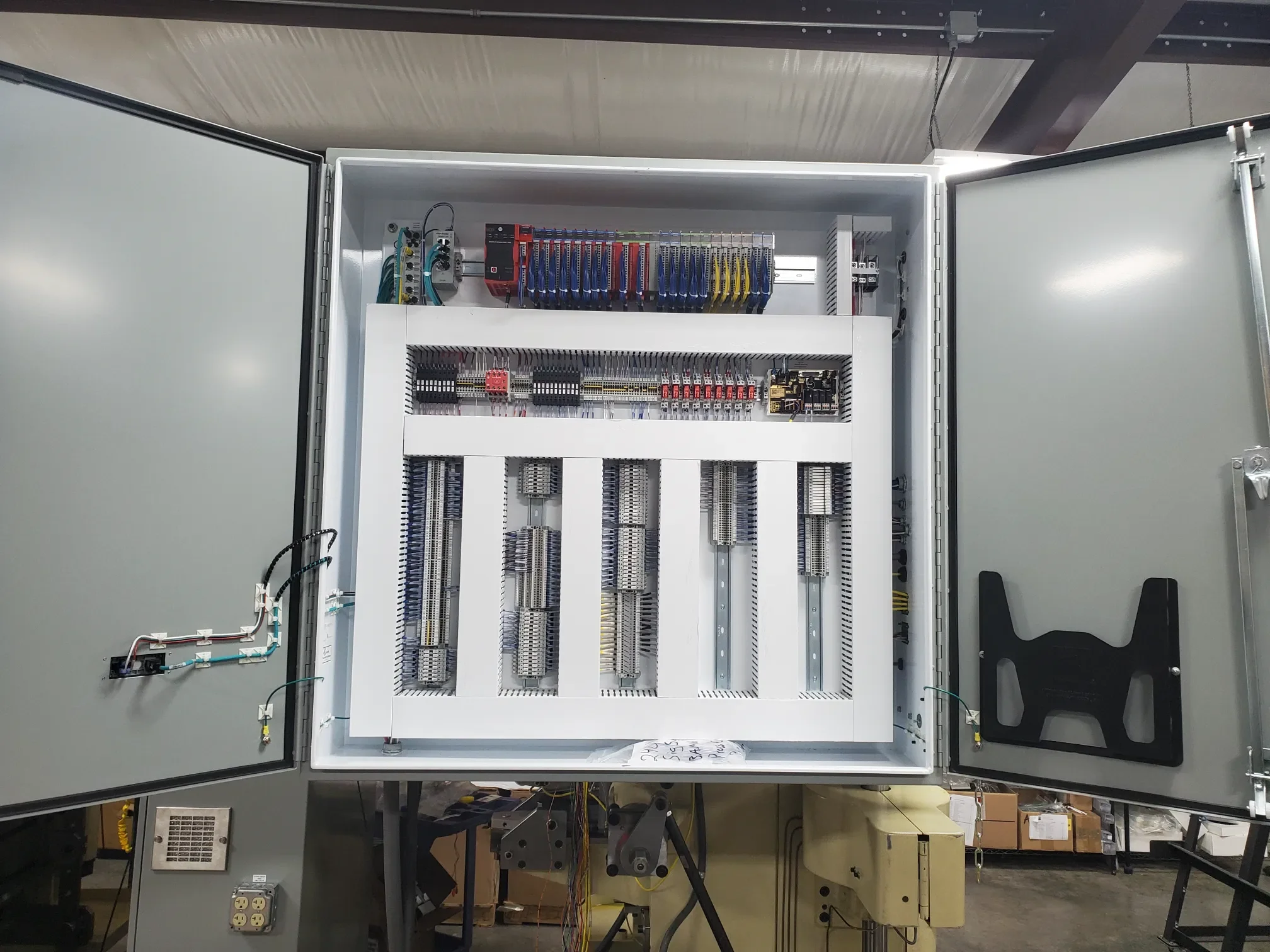 An open electrical control panel with various wires, circuit boards, and components inside, mounted on a gray metal cabinet, located in an industrial setting.