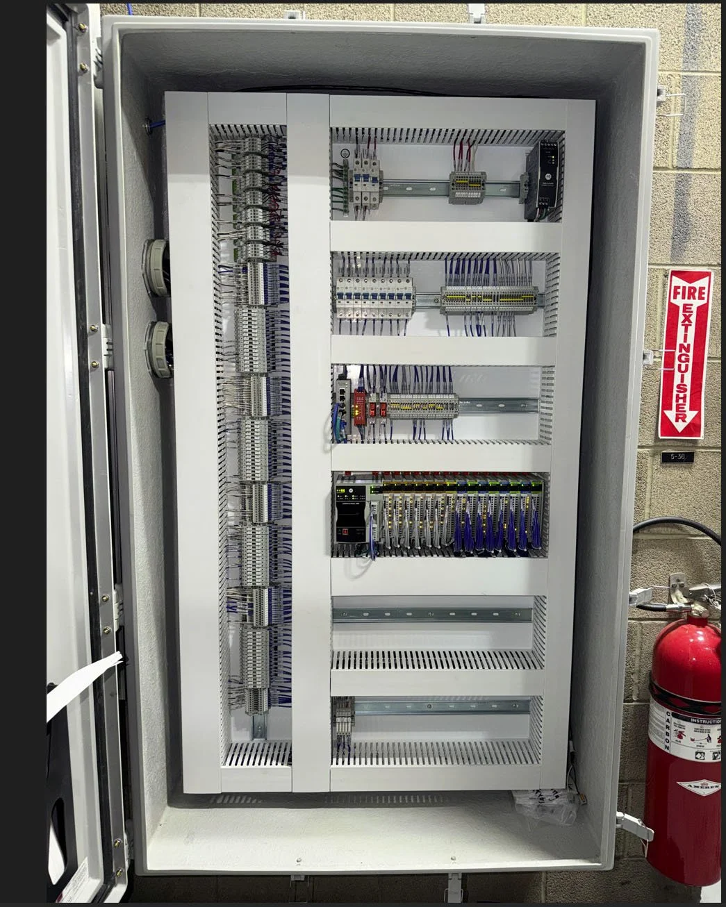 Electric control panel with various wiring and components inside a metal cabinet, mounted on a wall with a fire extinguisher nearby.