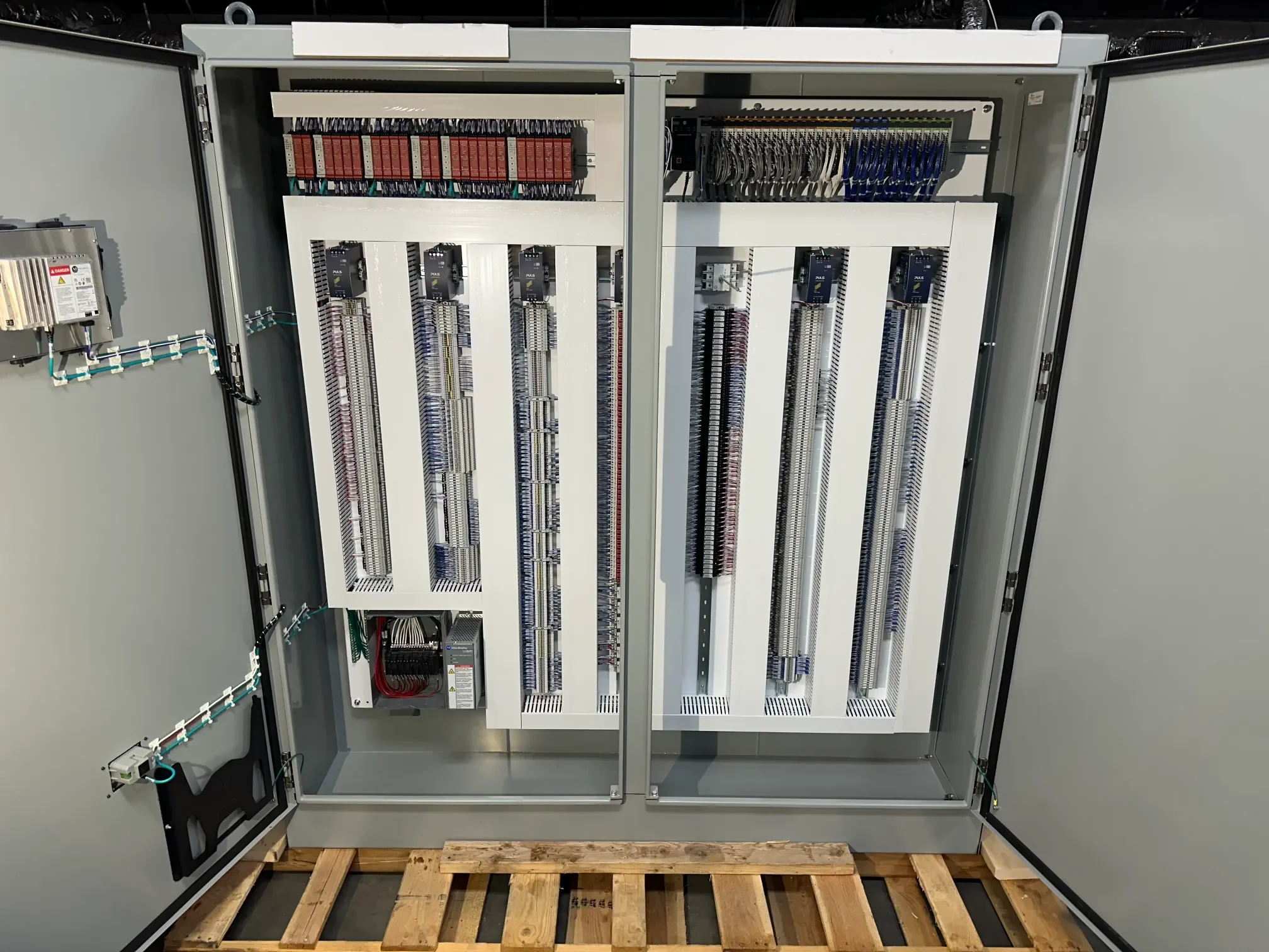 Open electrical control panel with various circuit boards, wiring, and components inside, mounted on a wooden pallet.