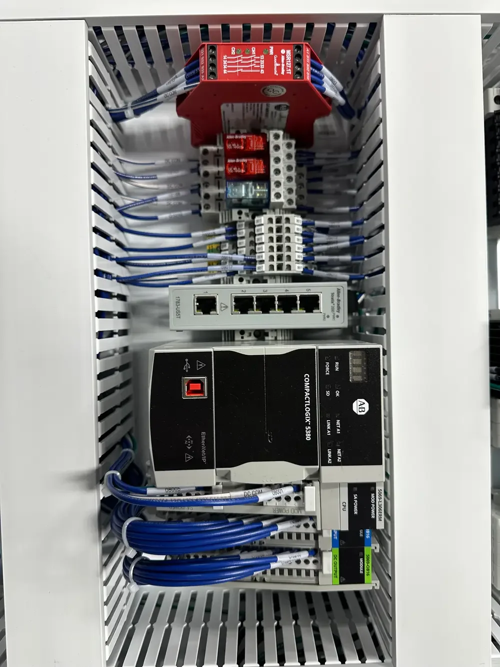 Inside an electrical control panel with various interconnected components including a red circuit board, a relay, Ethernet switches, and wiring.