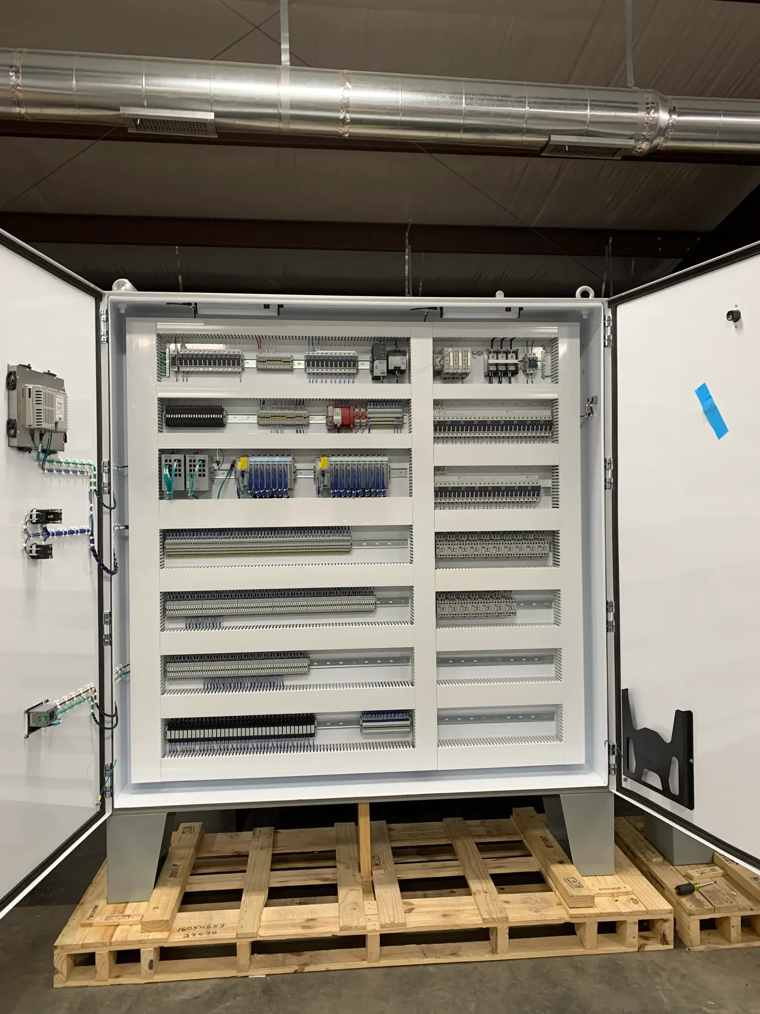 An open electrical control panel or circuit breaker box mounted on a white wall, set on a wooden pallet, with various wires, relays, and terminal blocks inside.