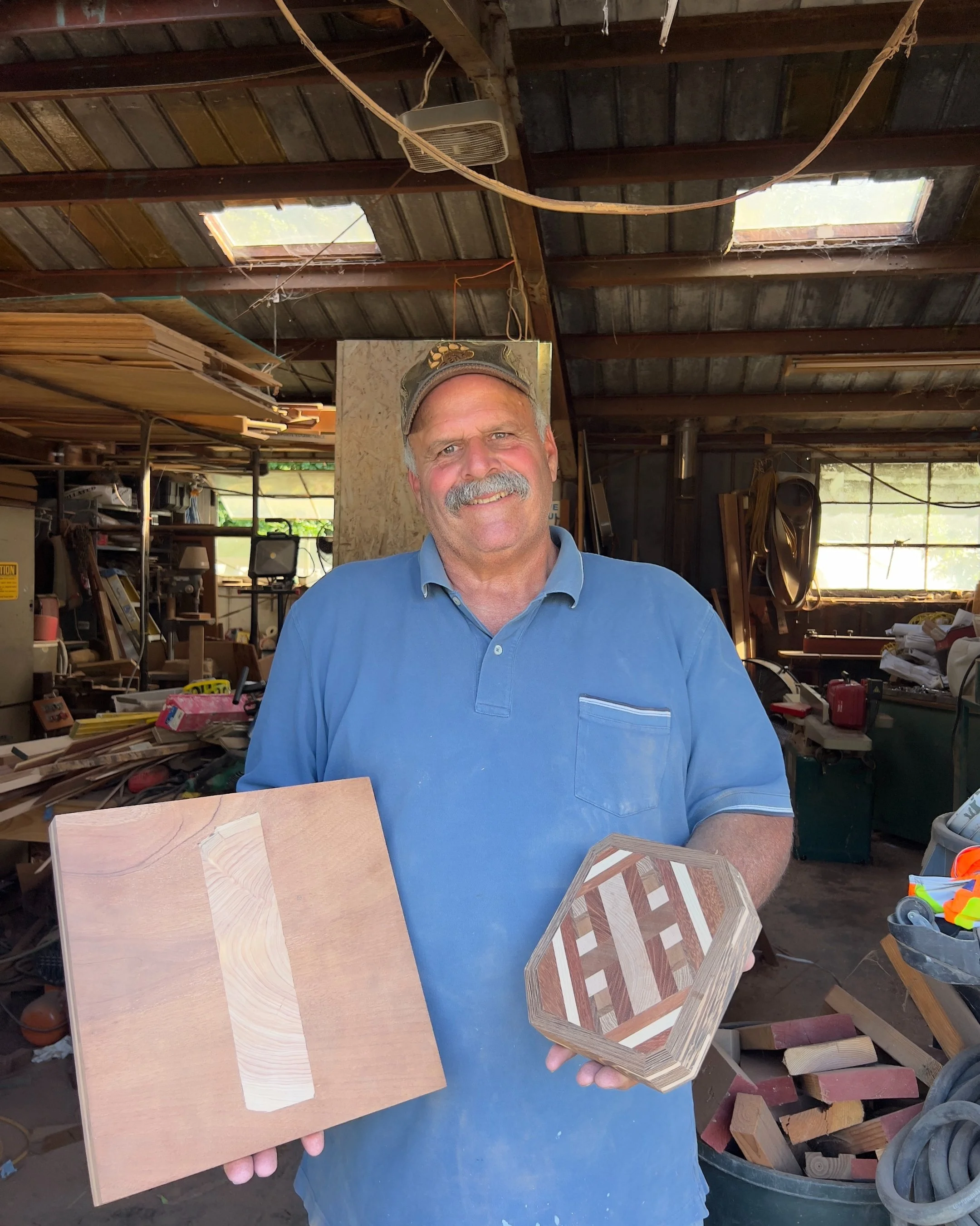 A smiling man with a mustache, wearing a blue shirt and cap, standing in a woodworking workshop holding a small octagon-shaped wooden piece and a square wooden board with a strip of lighter wood in the center.
