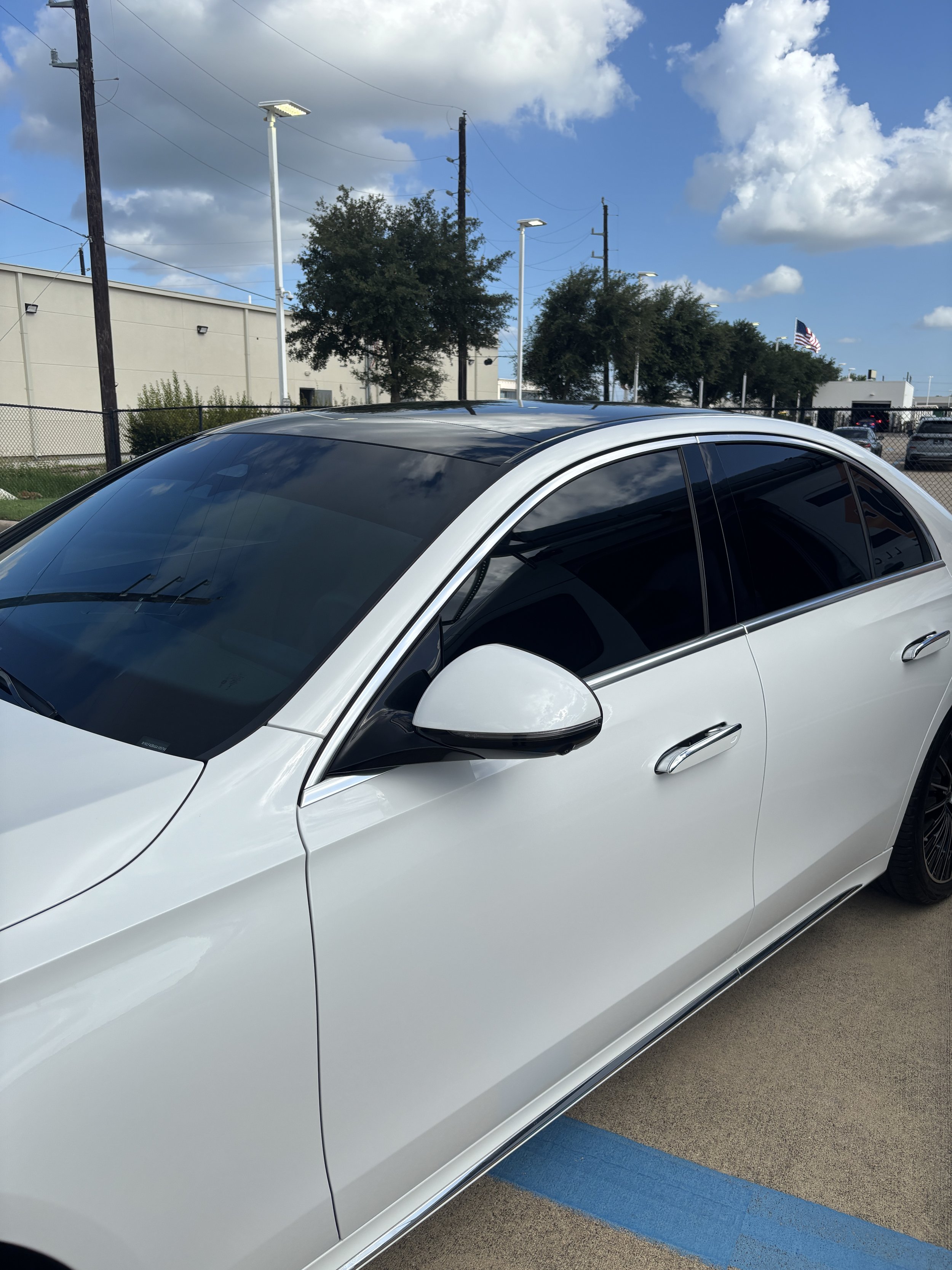A white Mercedes Benz on an open parking lot with trees and blue cloudy sky.
