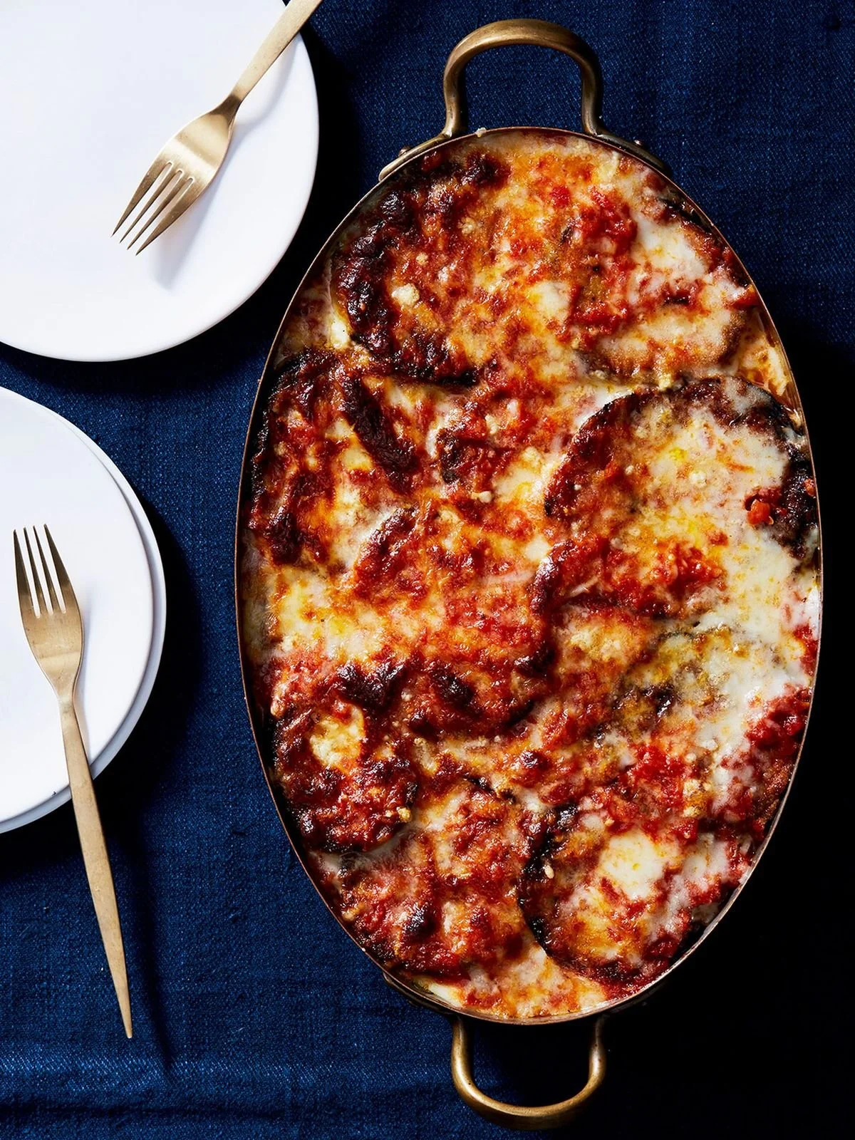 A baked eggplant Parmesan dish in an oval metal serving dish, with two place settings each with a white plate and a gold fork.