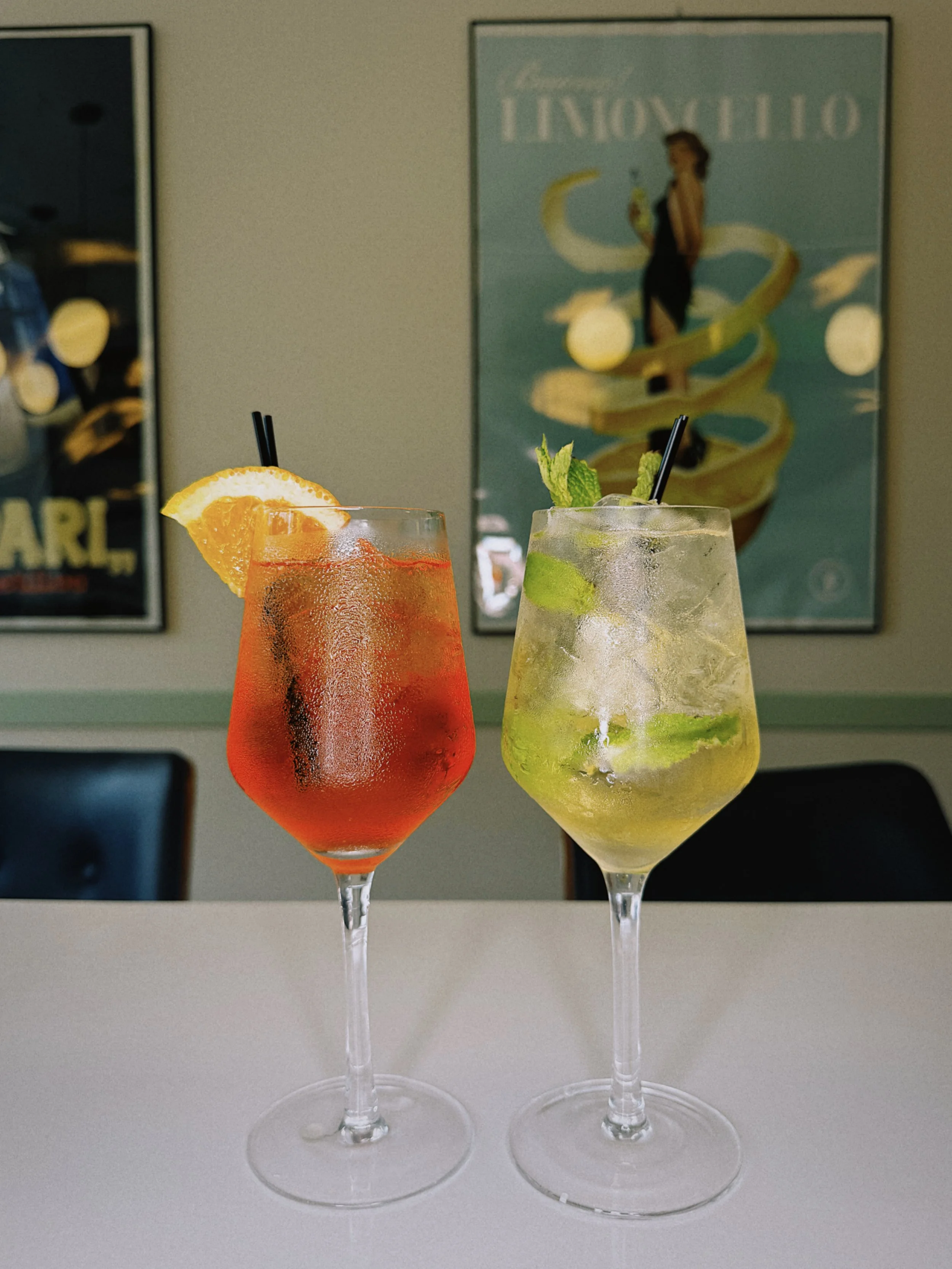 Two colorful cocktails in tall glasses on a white surface with a wall decorated with vintage posters in the background. One cocktail is red with an orange slice, and the other is yellow-green with mint leaves.