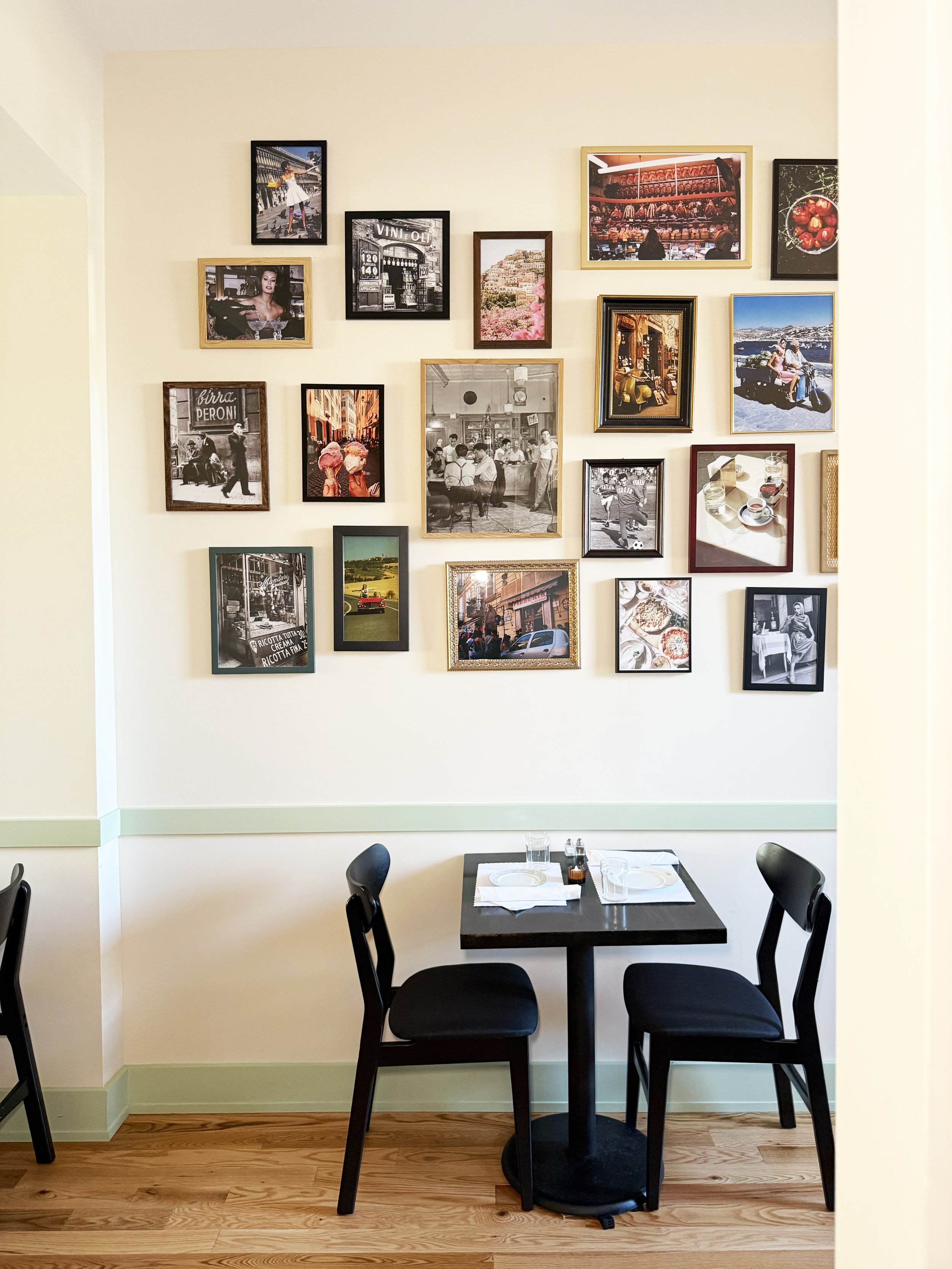 A small dining table set for two in a restaurant with black chairs, white plates, glasses, and utensils. The wall behind features a collage of colorful framed photographs of various scenes and people.