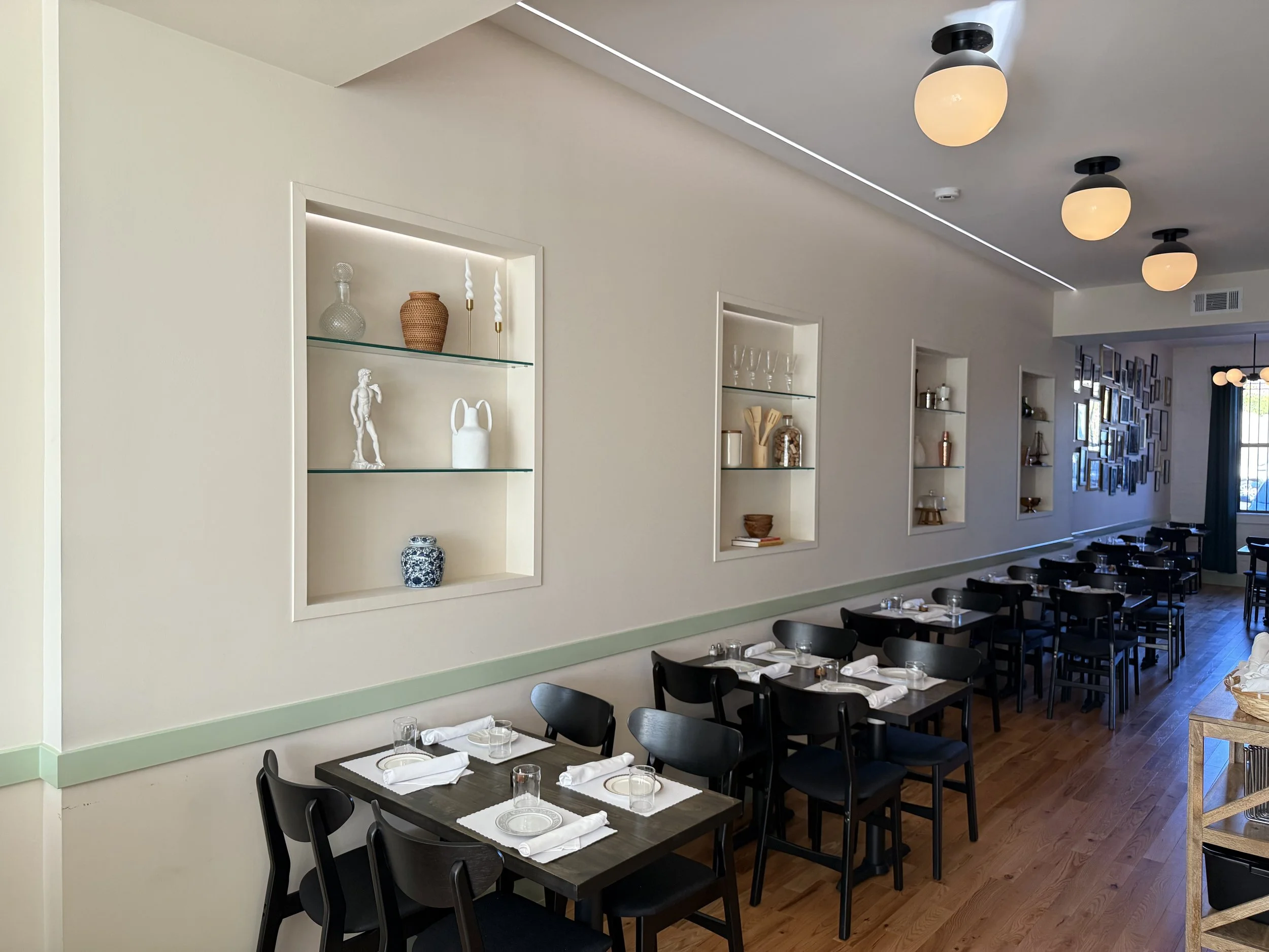Interior of a restaurant with black chairs and tables set with white napkins and glasses, featuring built-in wall shelves displaying decorative items, and a wall with photo frames and modern ceiling lights.