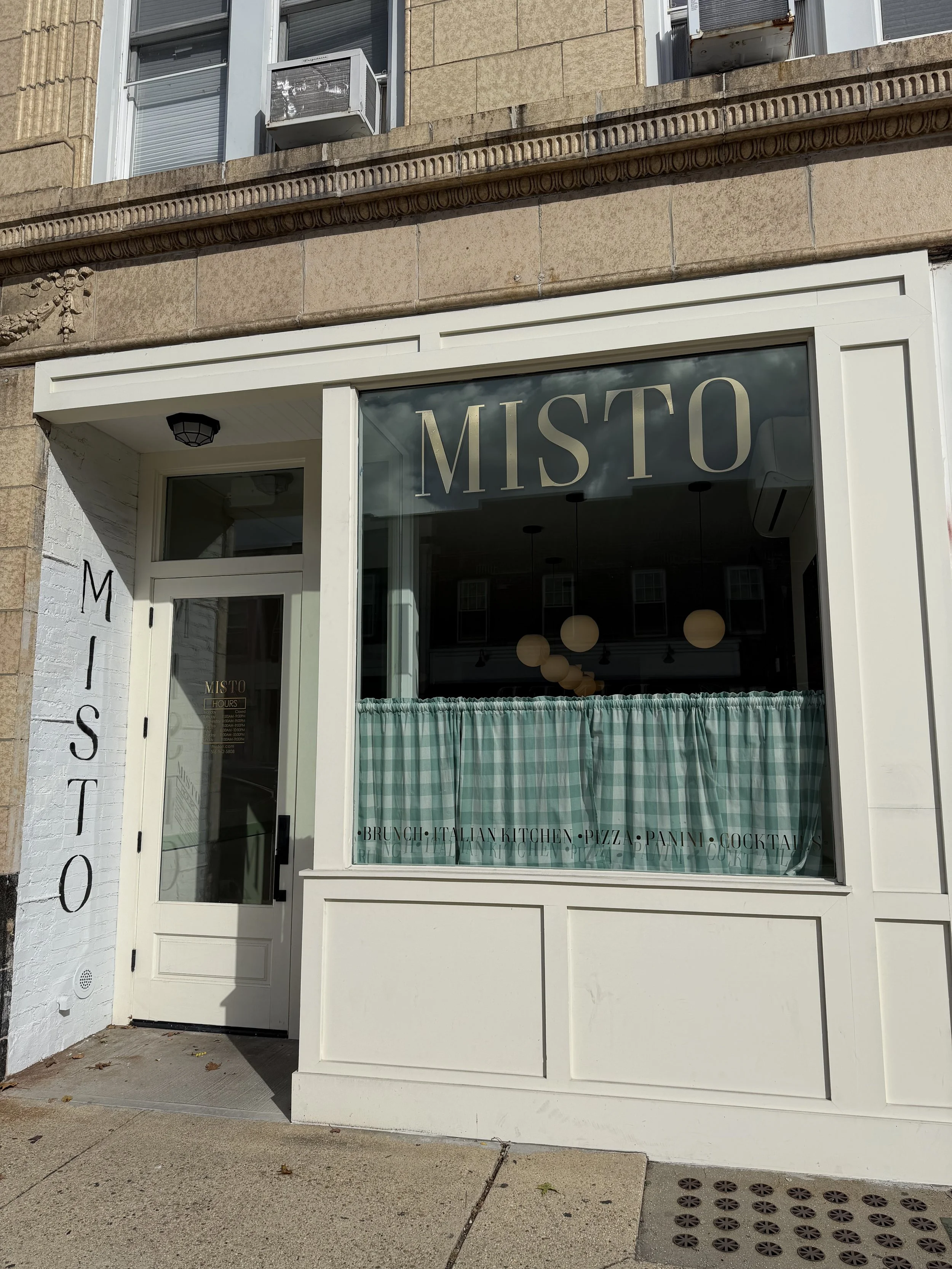 storefront for MISTO with large window and door, white exterior, green checkered curtains, and black letters spelling out 'MISTO' vertically on the brick wall next to the entrance.