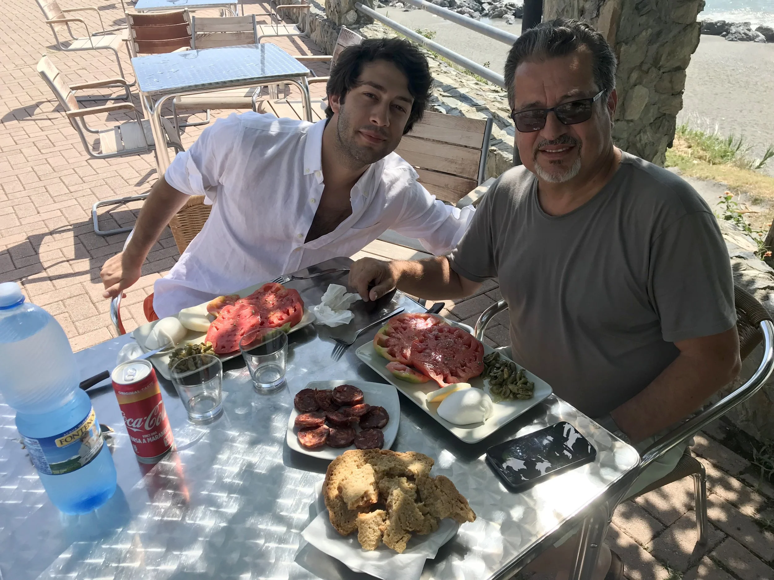 Two men sitting at an outdoor dining table with a meal that includes slices of watermelon, salami, condiments, bread, and a bottle of water and a soda. The man on the right is wearing glasses and a gray t-shirt, and the man on the left is wearing a white shirt. They are smiling and appear to be enjoying their meal, with a scenic background of rocky terrain and a beach.