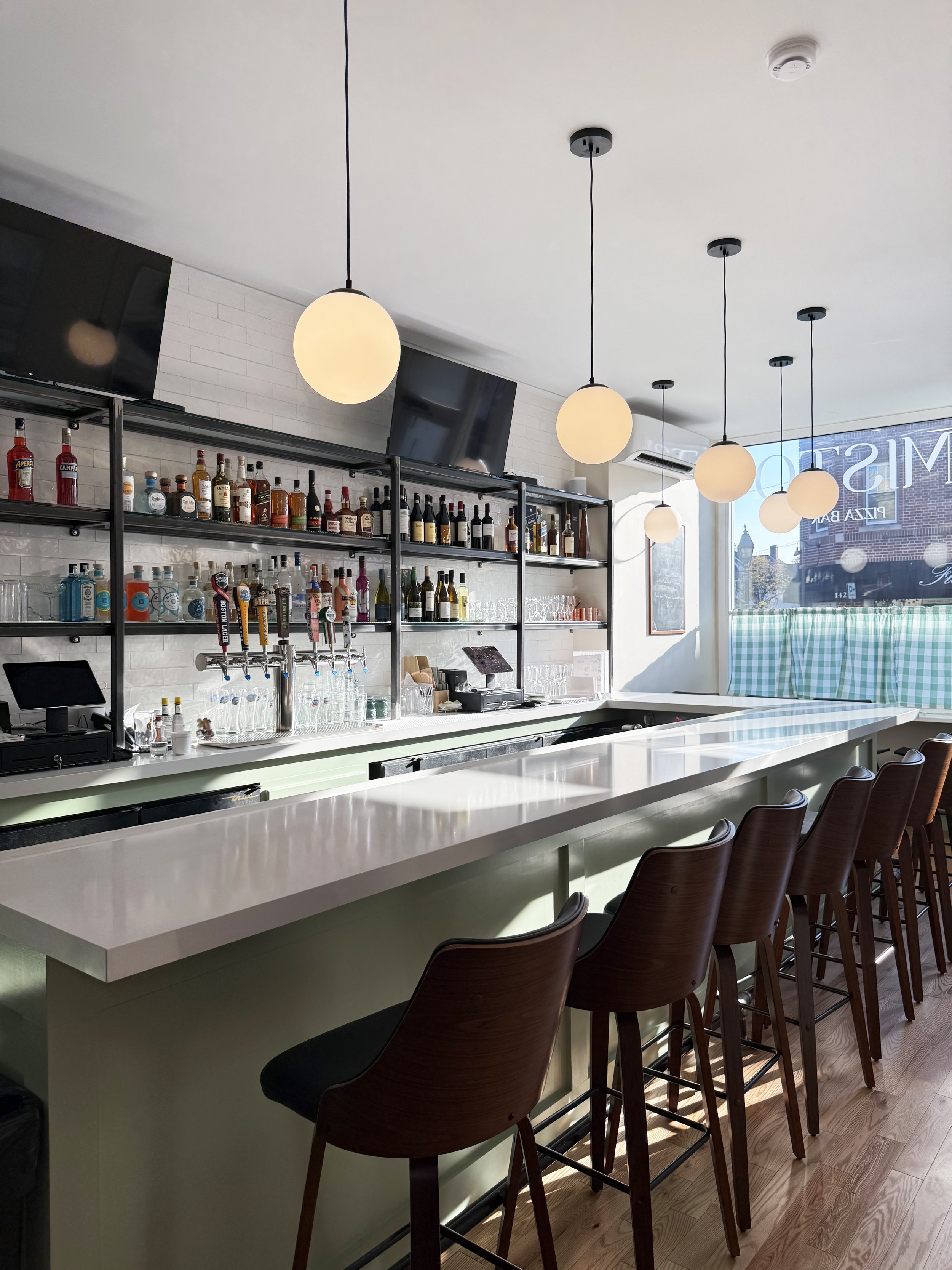 A bar with a white countertop, dark wooden bar stools, shelves with liquor bottles, televisions, and circular pendant lights hanging from the ceiling, with a window and greenery outside.