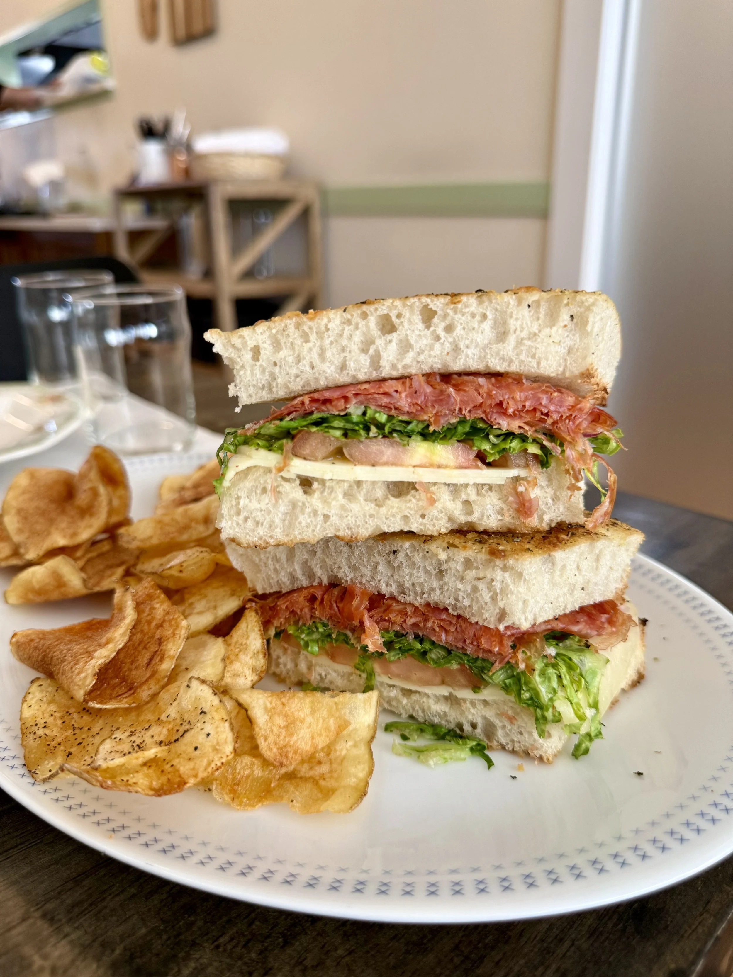 Close-up of a three-layer sandwich with beef, lettuce, and tomato on white bread, served with potato chips on a white plate.