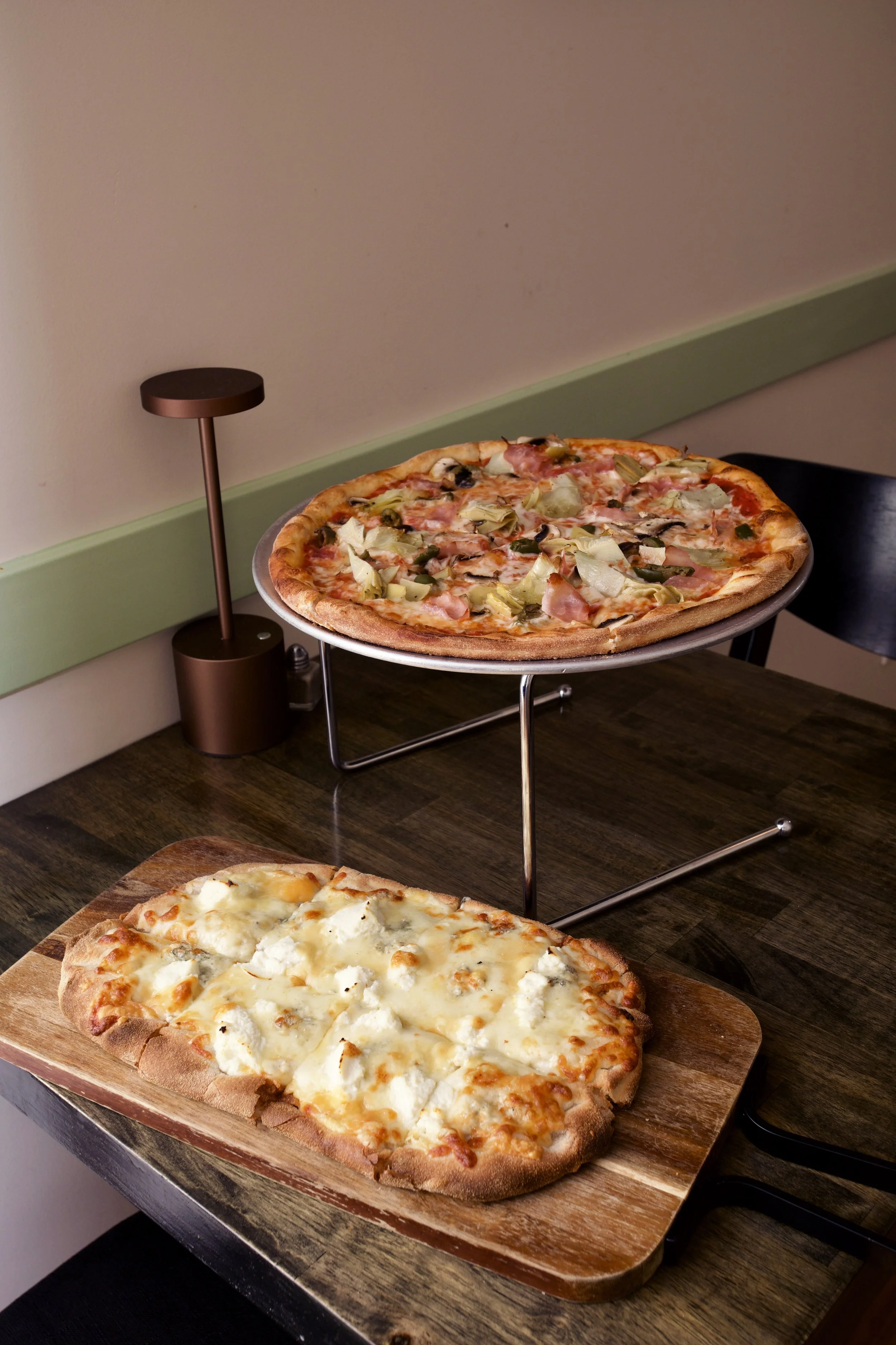 Two pizzas, one on a wooden serving board and one elevated on a stand, in a cozy indoor setting.
