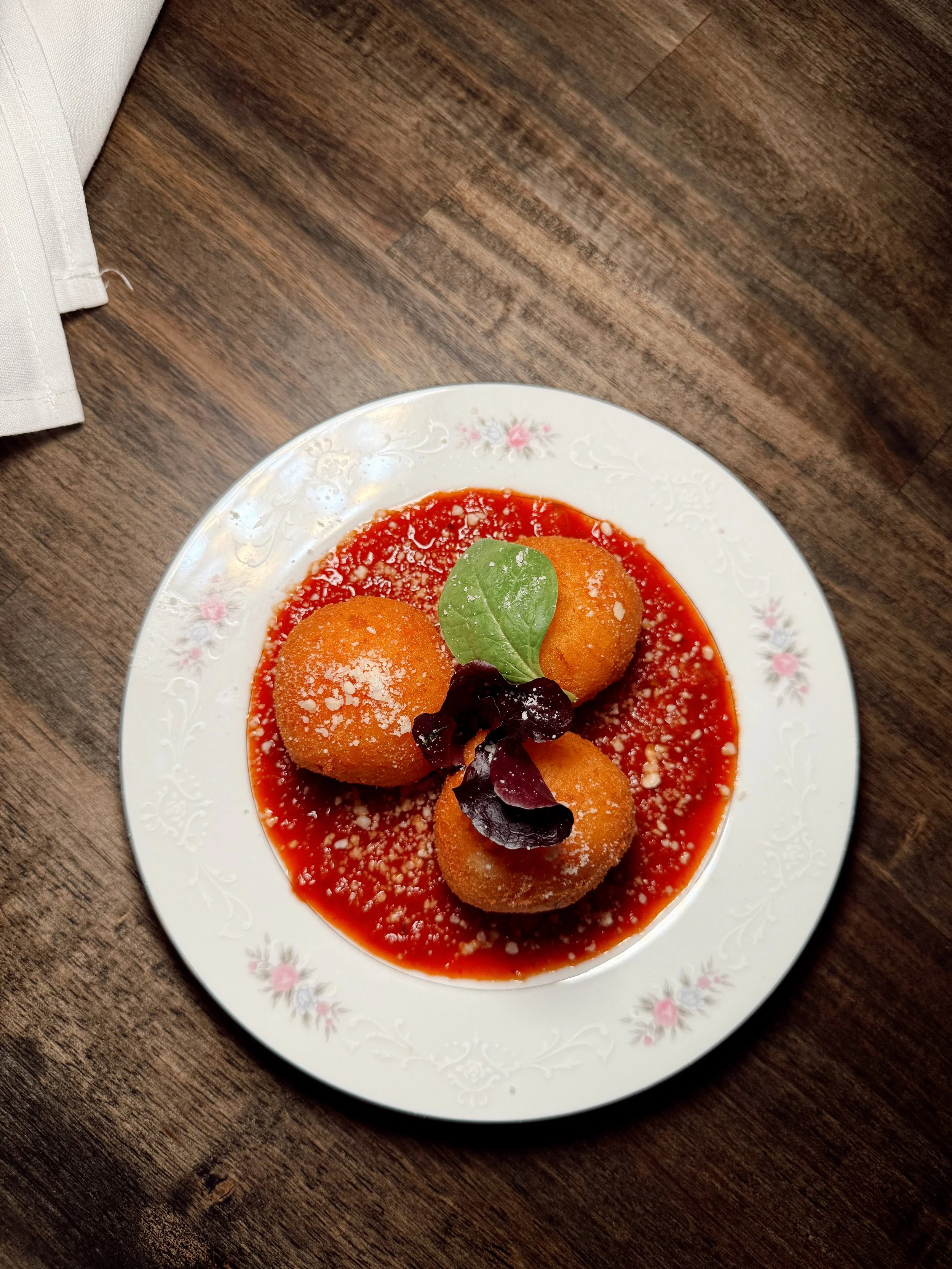 Three fried cheese balls topped with greenery and served on a floral-patterned plate with marinara sauce and grated cheese