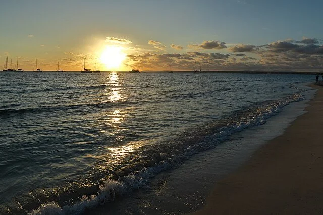 Sunset over the ocean with sailboats on the horizon and gentle waves hitting the sandy beach.
