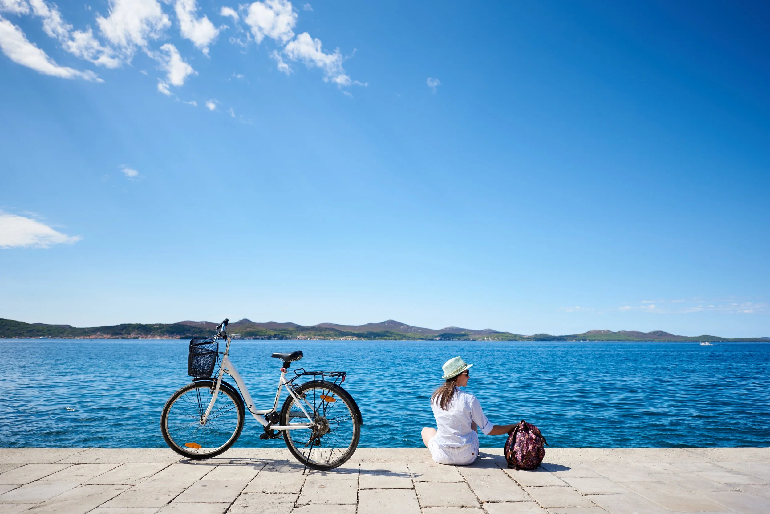 A woman sitting on a waterfront promenade with a bicycle nearby, looking at the water and distant hills under a clear blue sky.