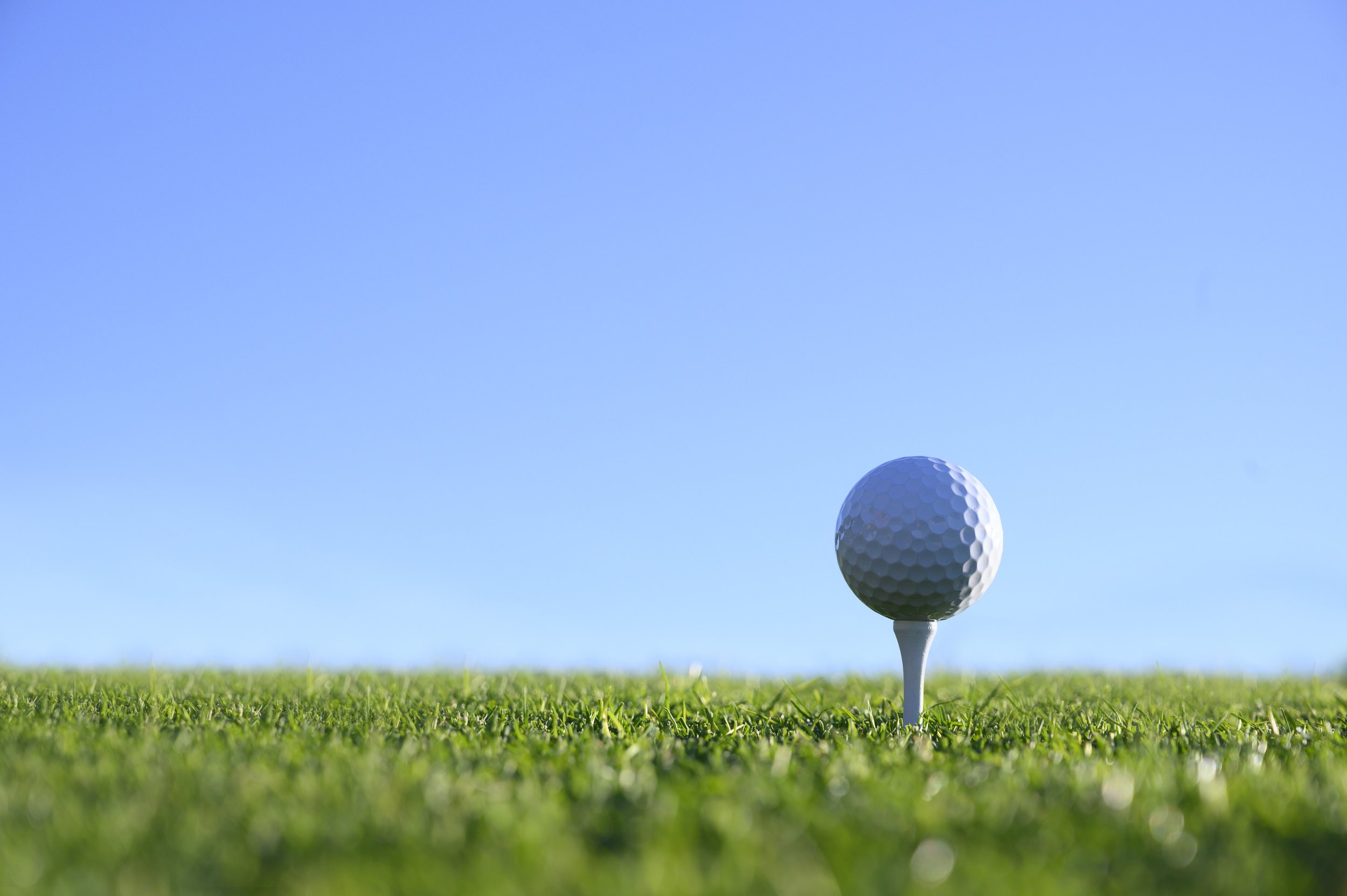 A golf ball on a tee ready for a drive on the green grass with a clear blue sky in the background.