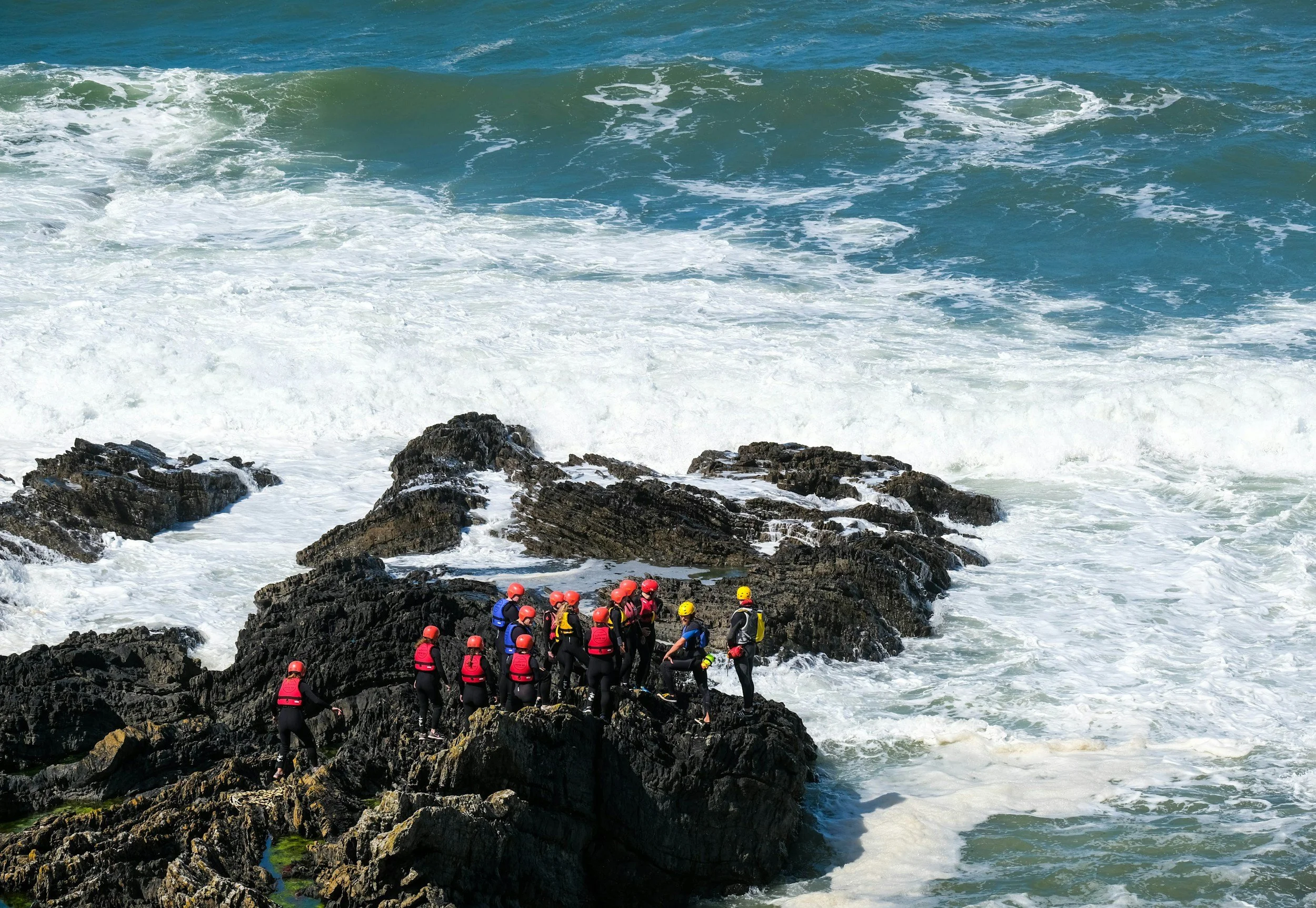 Group of people in safety gear gather on rocks near the ocean with waves crashing around them.