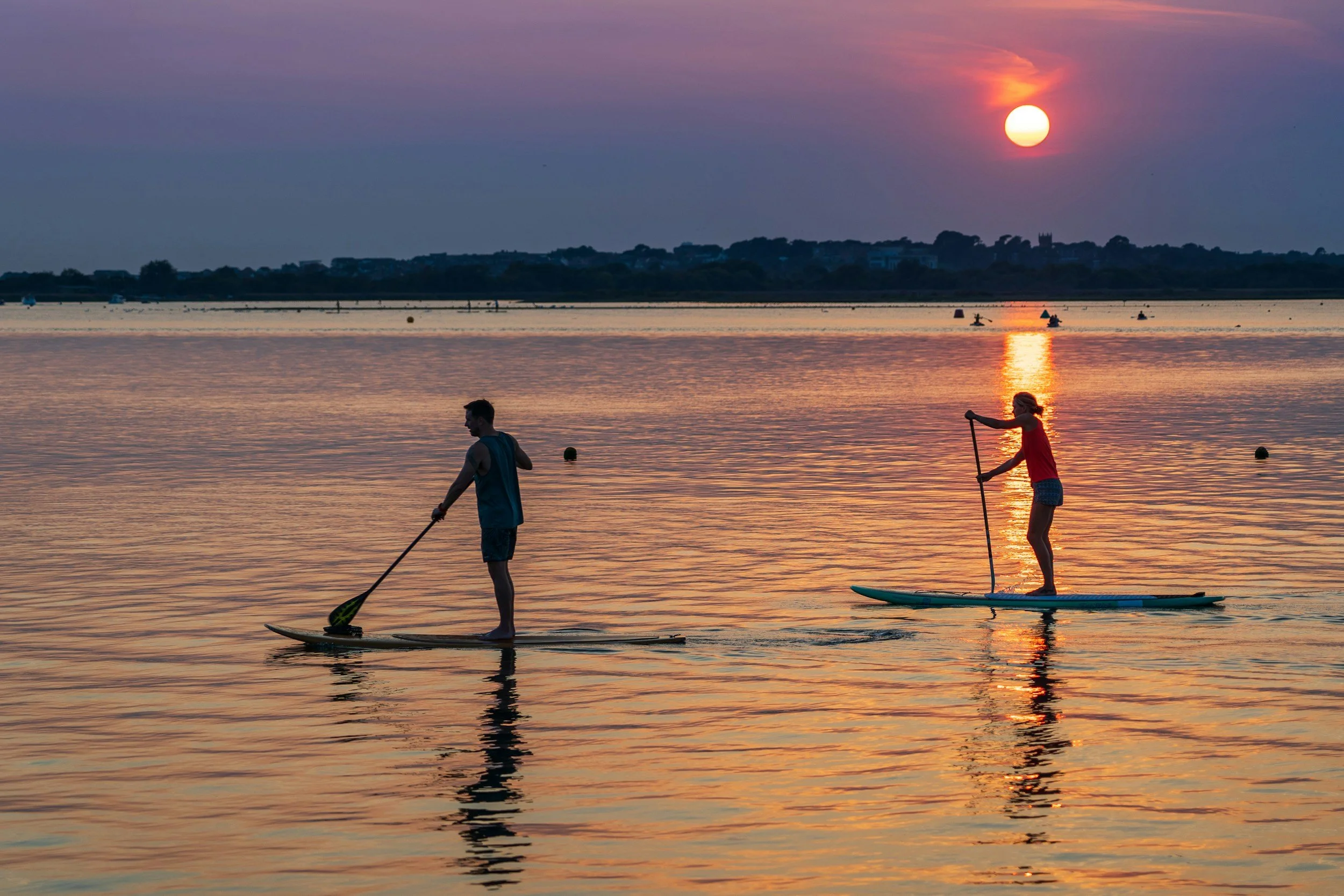 Two people paddleboarding on calm water during sunset, with the sun low in the sky casting reflections on the water, and boats visible in the distance.