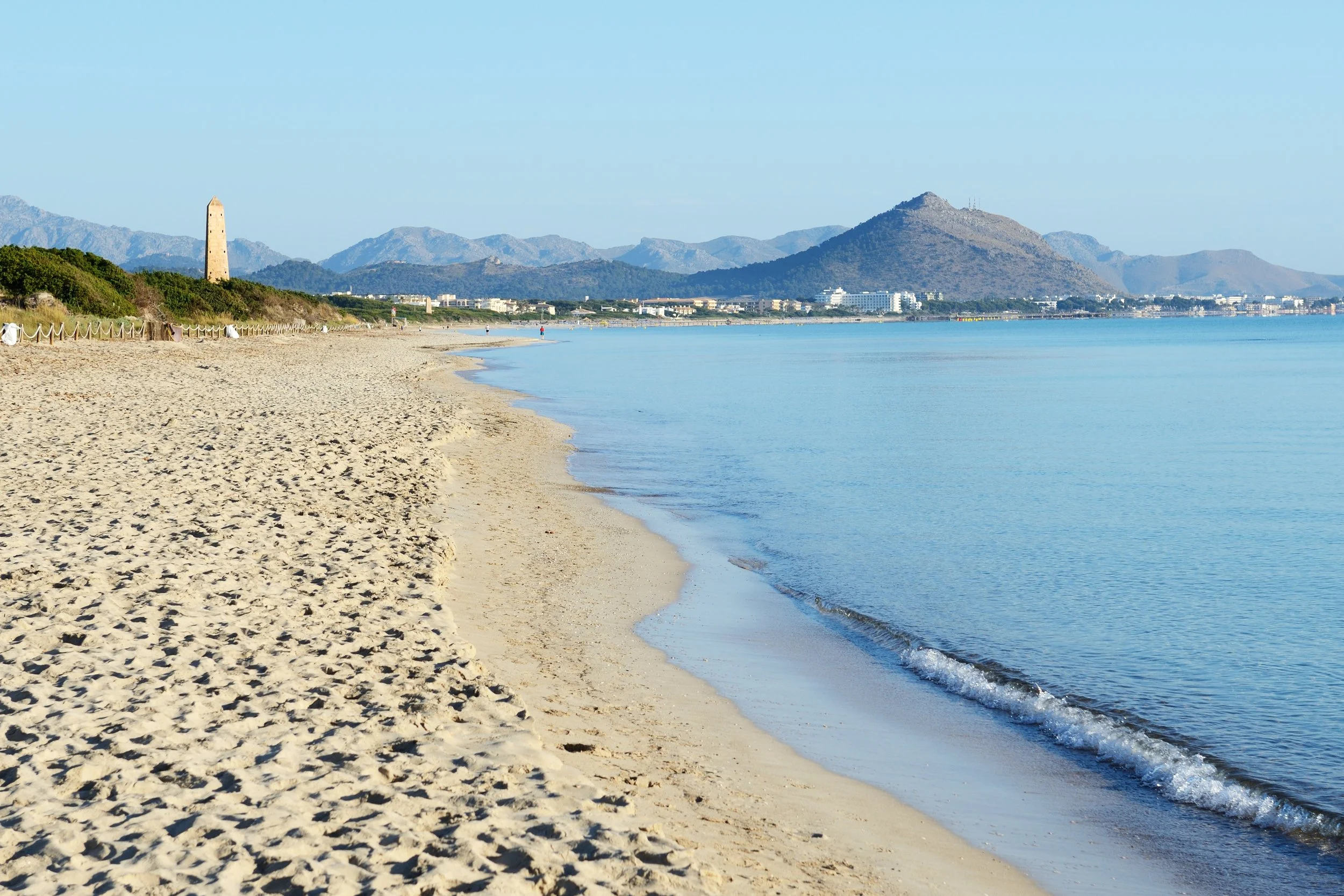 Scenic view of a calm beach with light-colored sand, clear blue water, distant mountains, and a clear sky.