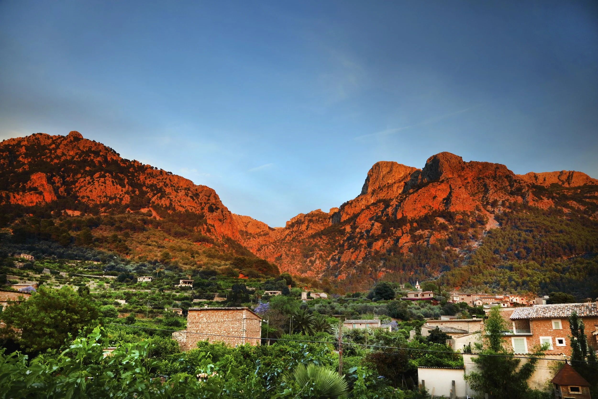 Sunset lighting on a mountain range with a village at the base, green trees and houses in the foreground.